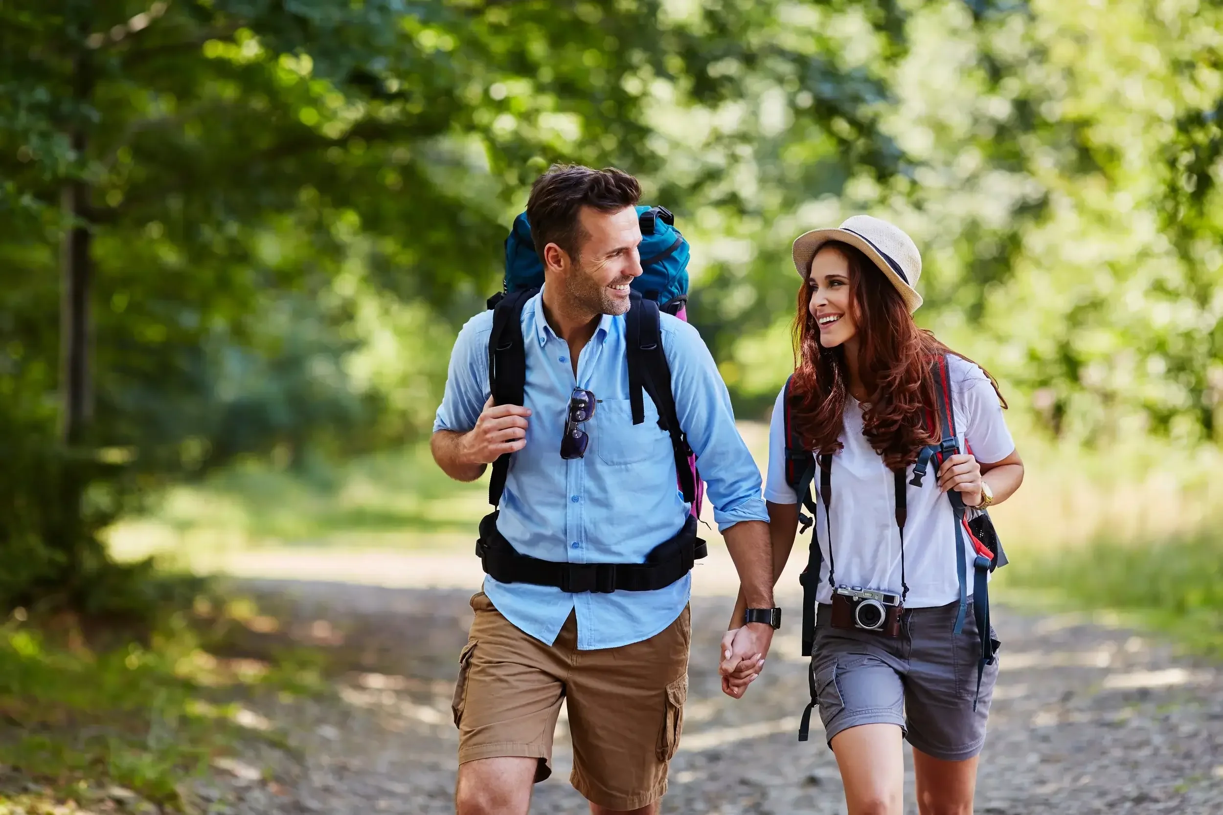 A smiling man and woman hiking hand-in-hand through a wooded trail, both carrying backpacks and wearing casual outdoor clothes and hats.