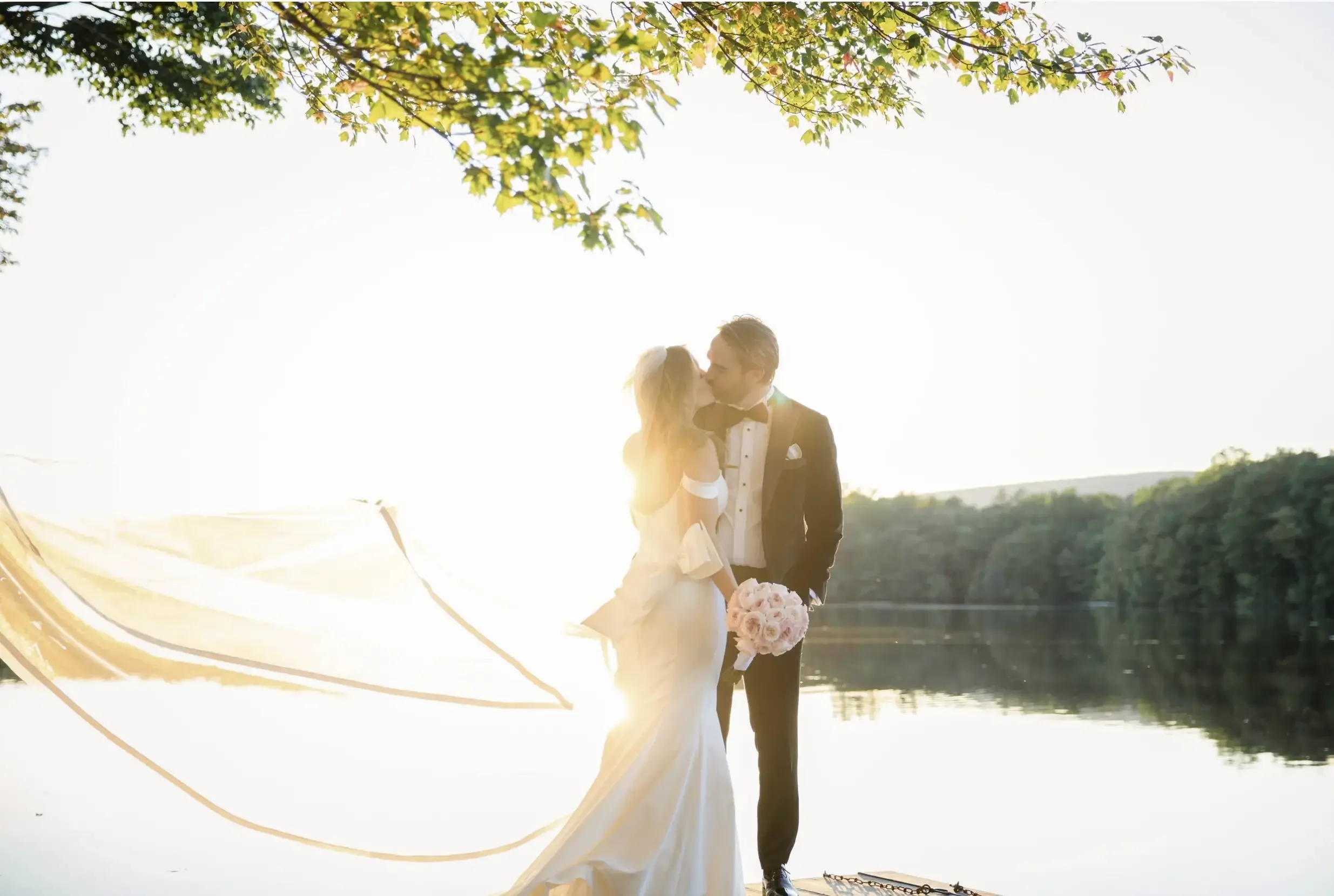 A bride and groom sharing a kiss by a river during sunset, with the bride holding a bouquet of pink roses and the groom wearing a tuxedo.