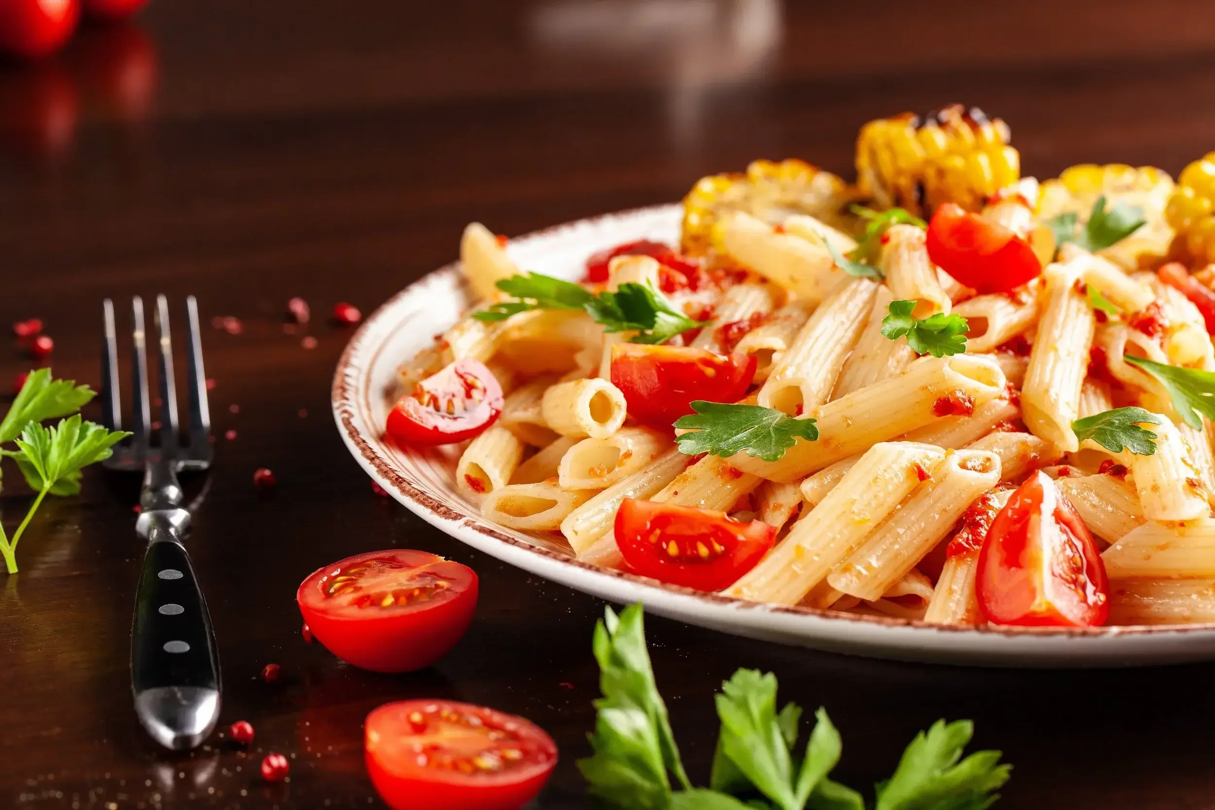 Plate of cooked penne pasta with cherry tomatoes and parsley, garnished with corn, served on a dark wooden table