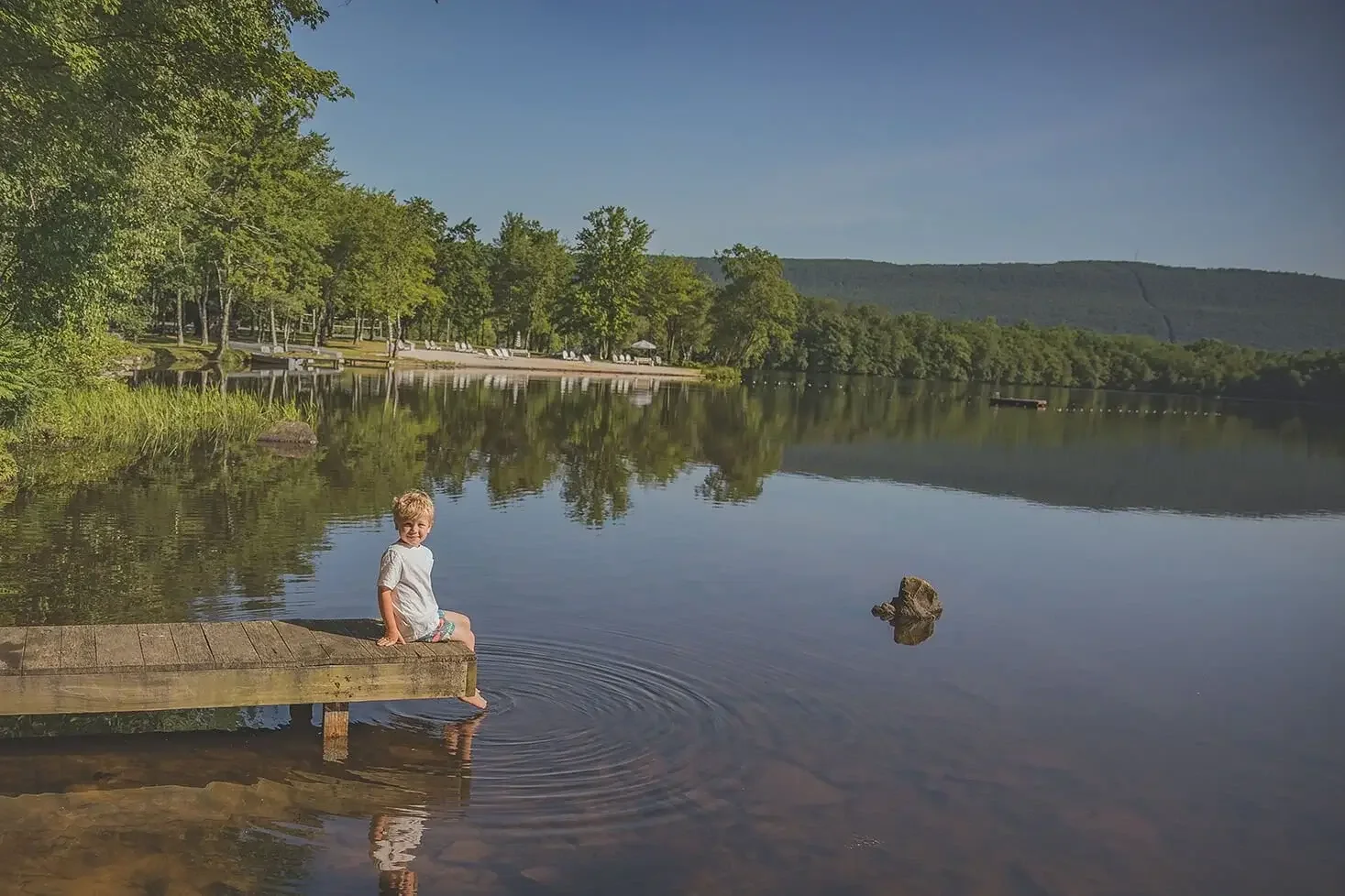 A young boy sitting on a wooden dock by the lake, smiling, with trees and a shoreline in the background on a sunny day.