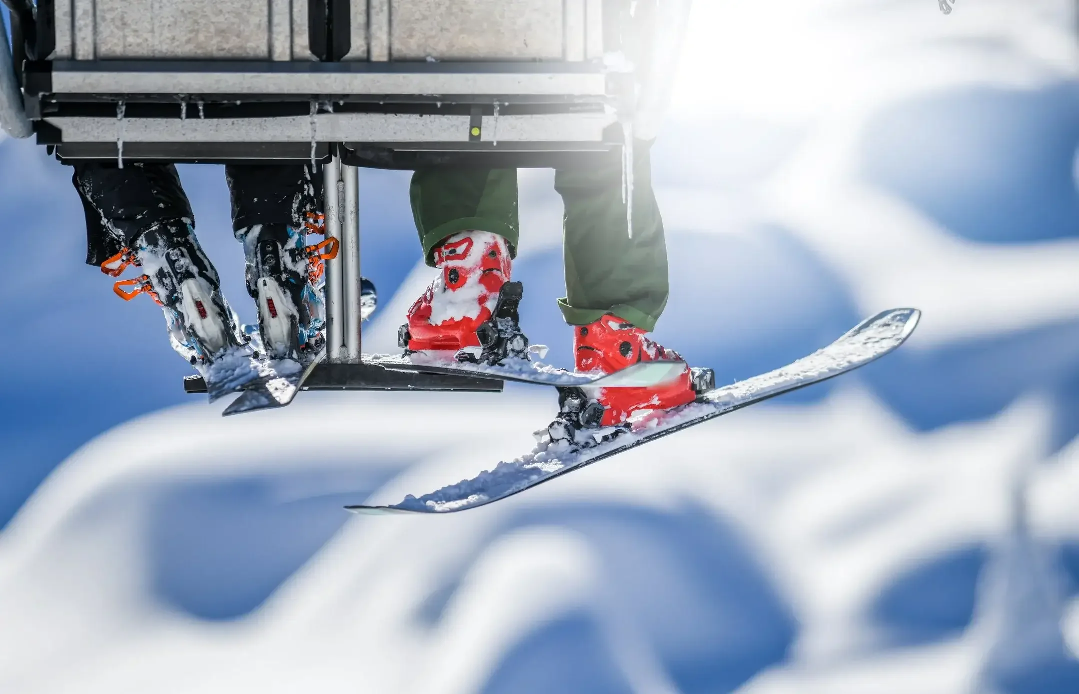 Close-up of a skier's feet on a ski lift, wearing red ski boots, with snow and a clear blue sky in the background.
