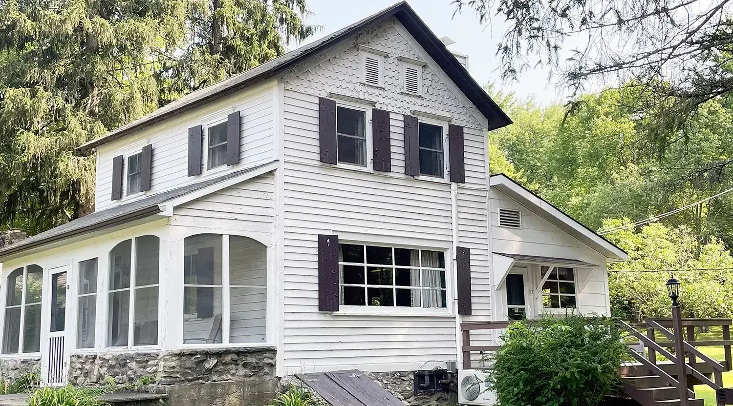 A two-story white house with black shutters and a stone foundation, surrounded by green trees and a well-kept lawn.
