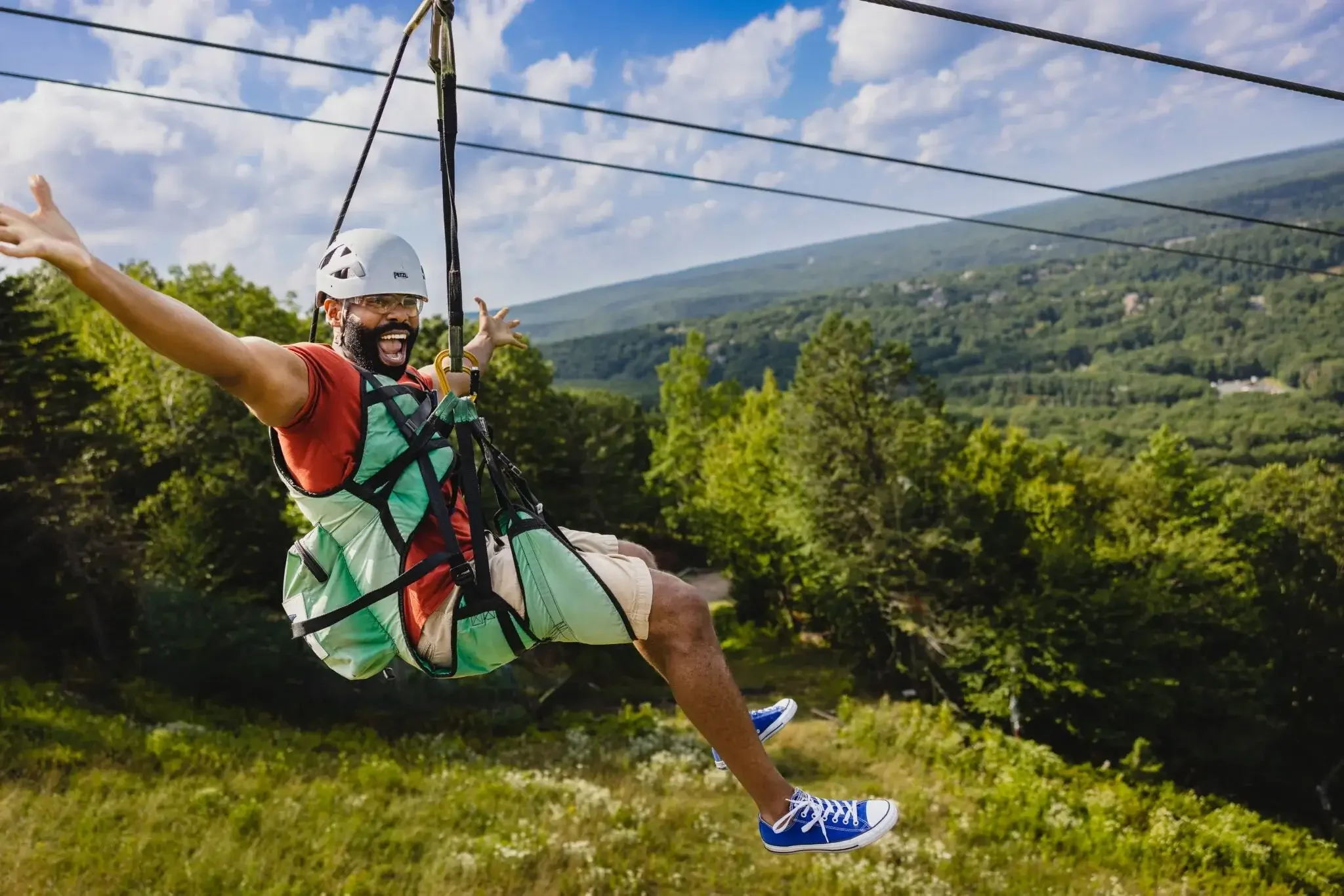 Man on zip line with arms outstretched, smiling, wearing helmet, harness, red shirt, beige shorts, and blue sneakers, flying over a lush green landscape with trees and mountains in the background.