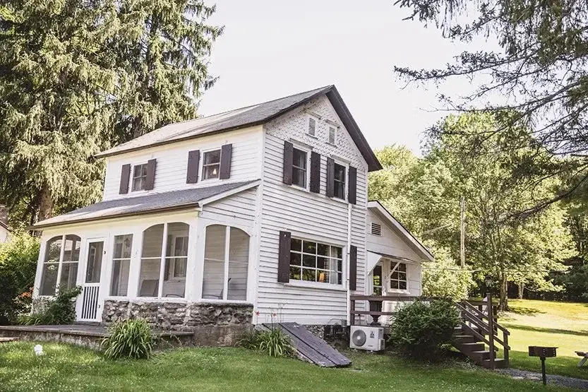 Two-story white house with black shutters, a porch, and stairs leading to a grassy yard surrounded by trees.