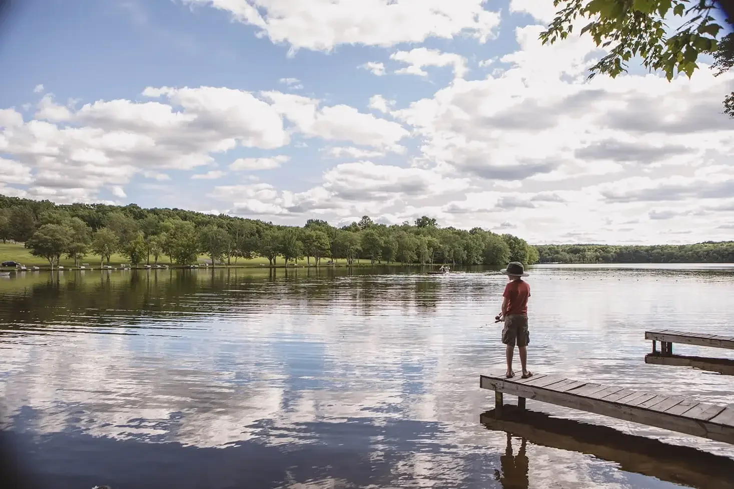 A person standing on a dock fishing in a calm lake under a partly cloudy sky, with trees lining the distant shoreline.