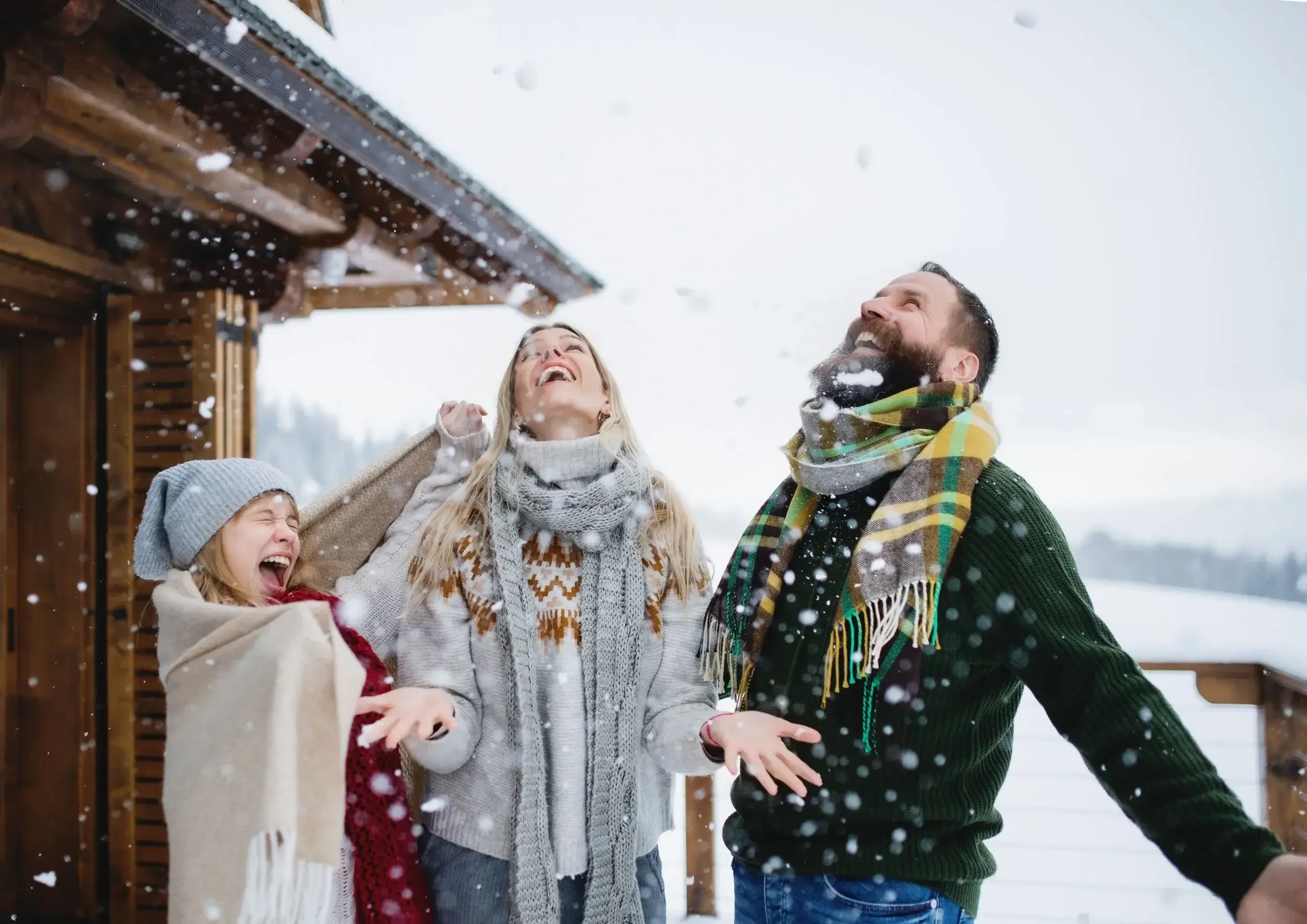 A family enjoying winter outside, throwing snow in the air and laughing, standing on a snow-covered deck near a wooden house.