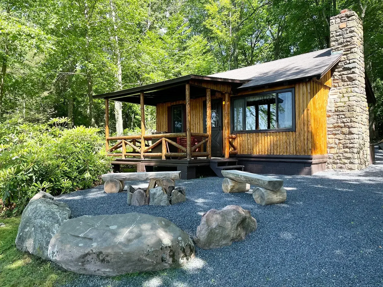 A small wooden cabin with a stone chimney, nestled in a wooded area with green trees, features a porch with a wooden railing and outdoor seating with rocks and logs, under a partly shaded sky.