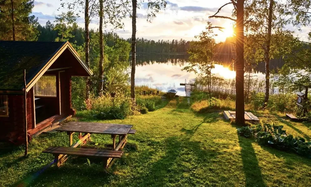 Wooden cabin by a calm forest lake at sunset.
