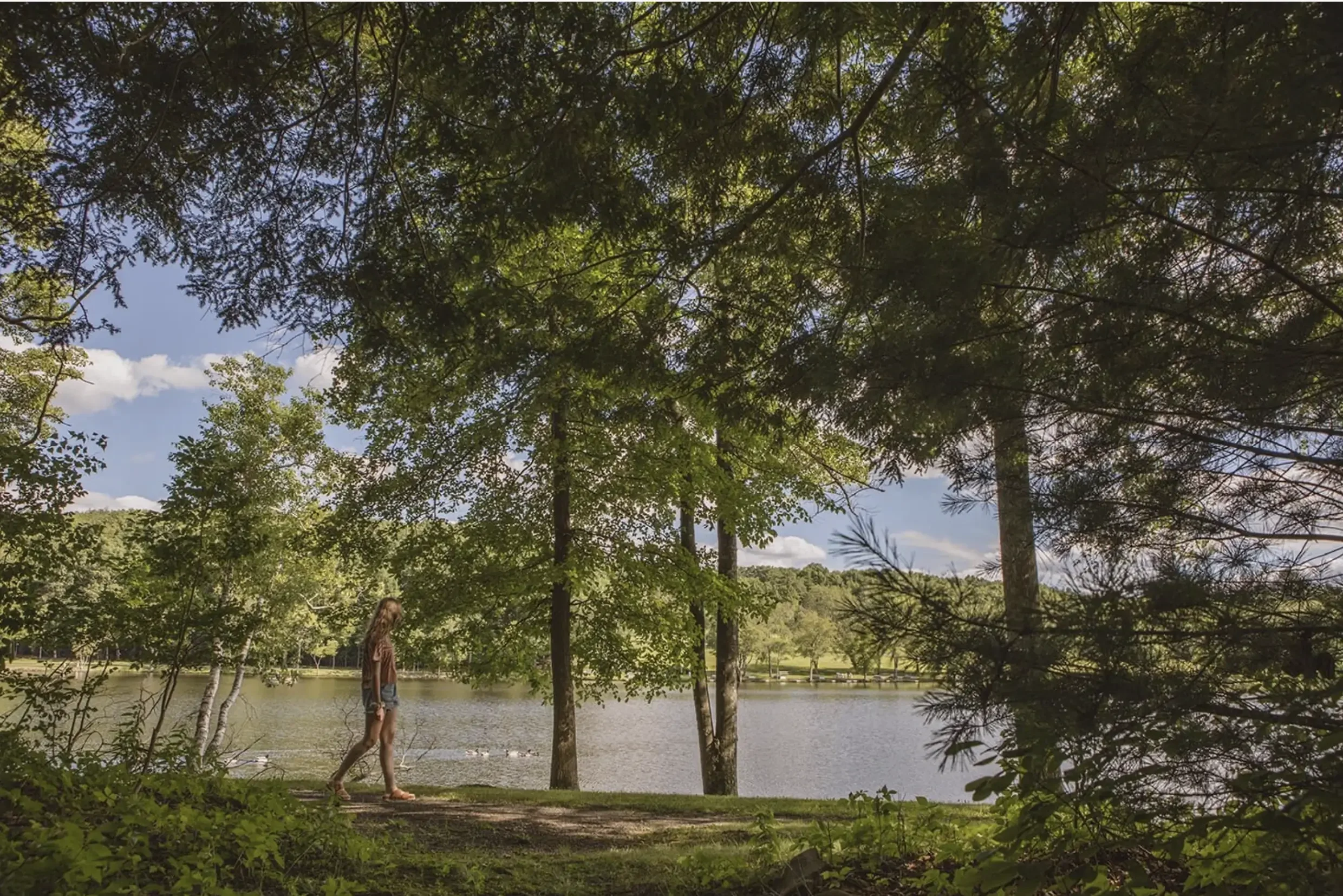 A woman walking along a wooded lakeside trail, with trees and water in the background under a partly cloudy sky.