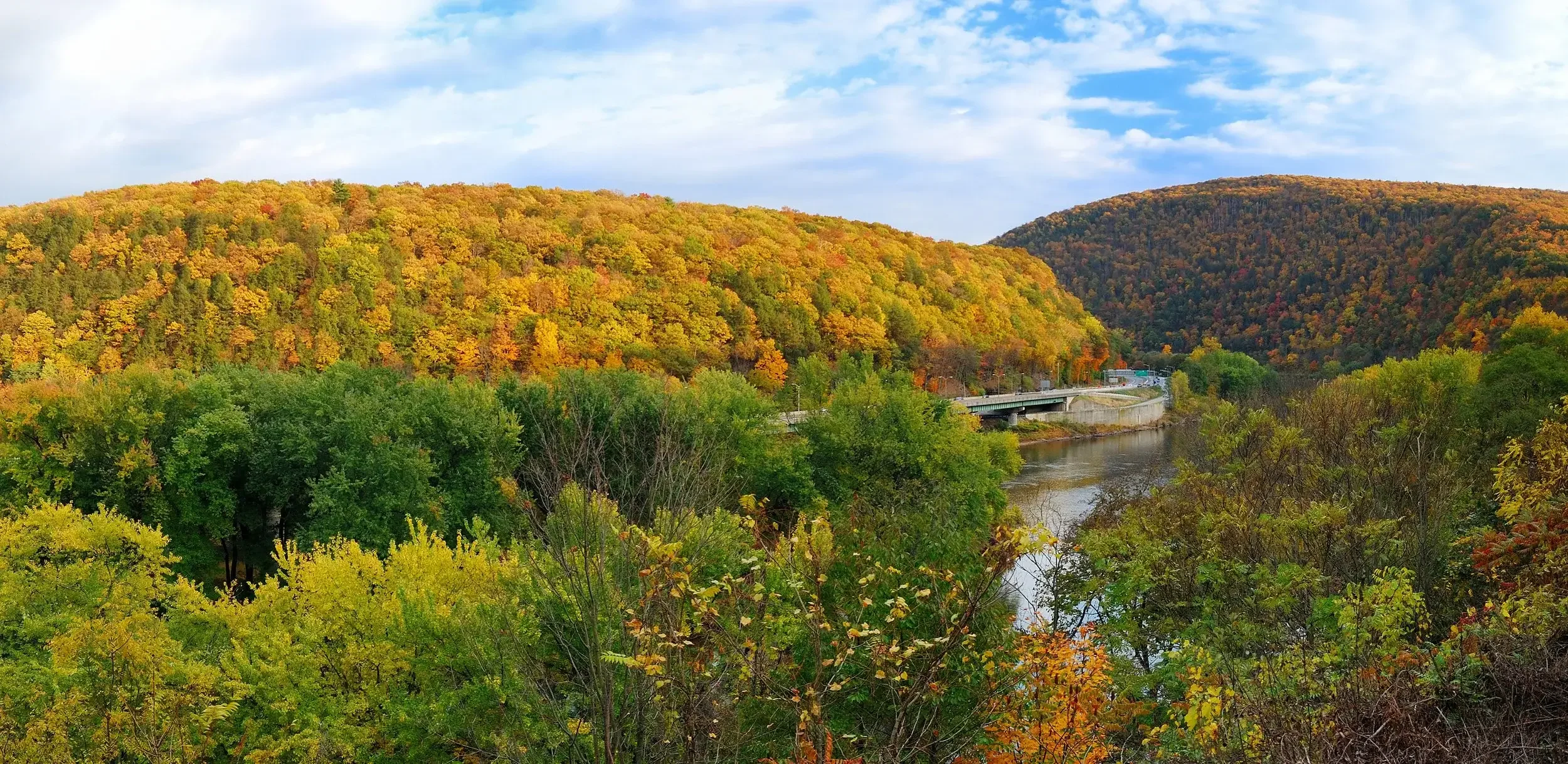 Colorful fall landscape with trees in green, yellow, orange, and red, a river, hills, and a cloudy sky.