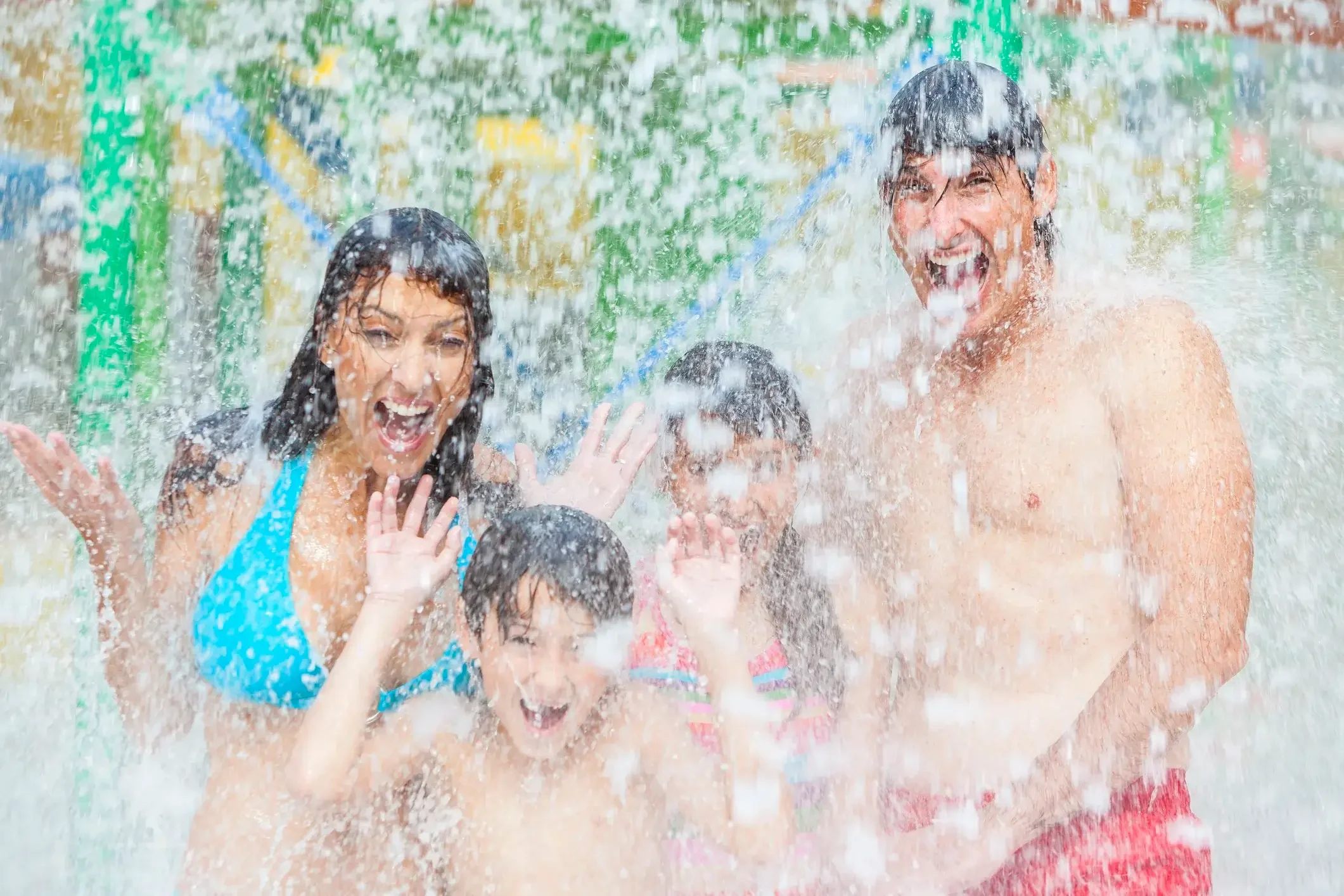 A family of four, two adults and two children, enjoying themselves under a water spray at a water park or splash pad. They are smiling and wearing swimsuits.