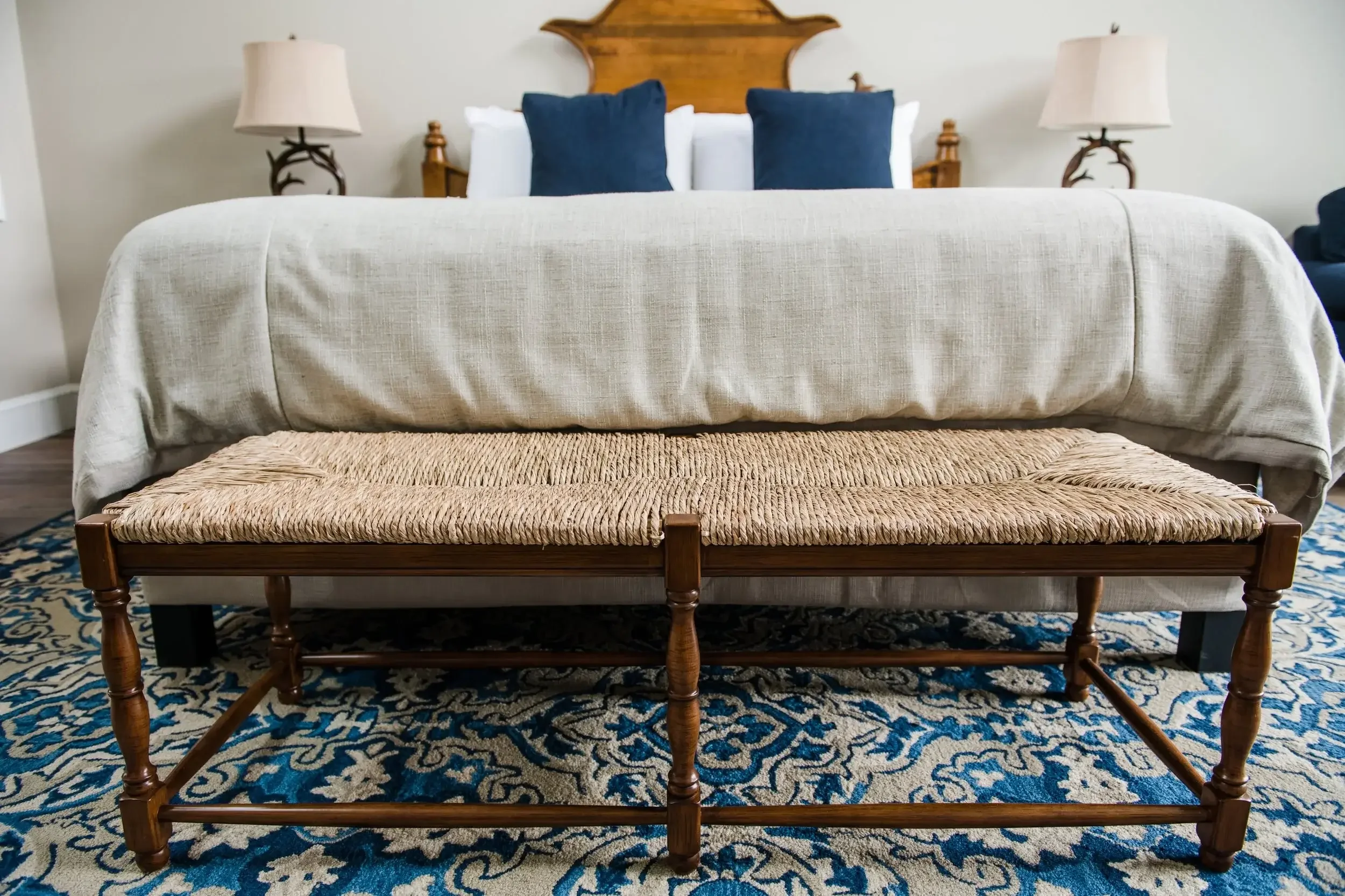 A bedroom with a large bed featuring a beige bedspread, two navy pillows, and a wooden headboard. There are matching side tables with beige lamps, and a woven bench at the foot of the bed. The room has a patterned blue and beige area rug.