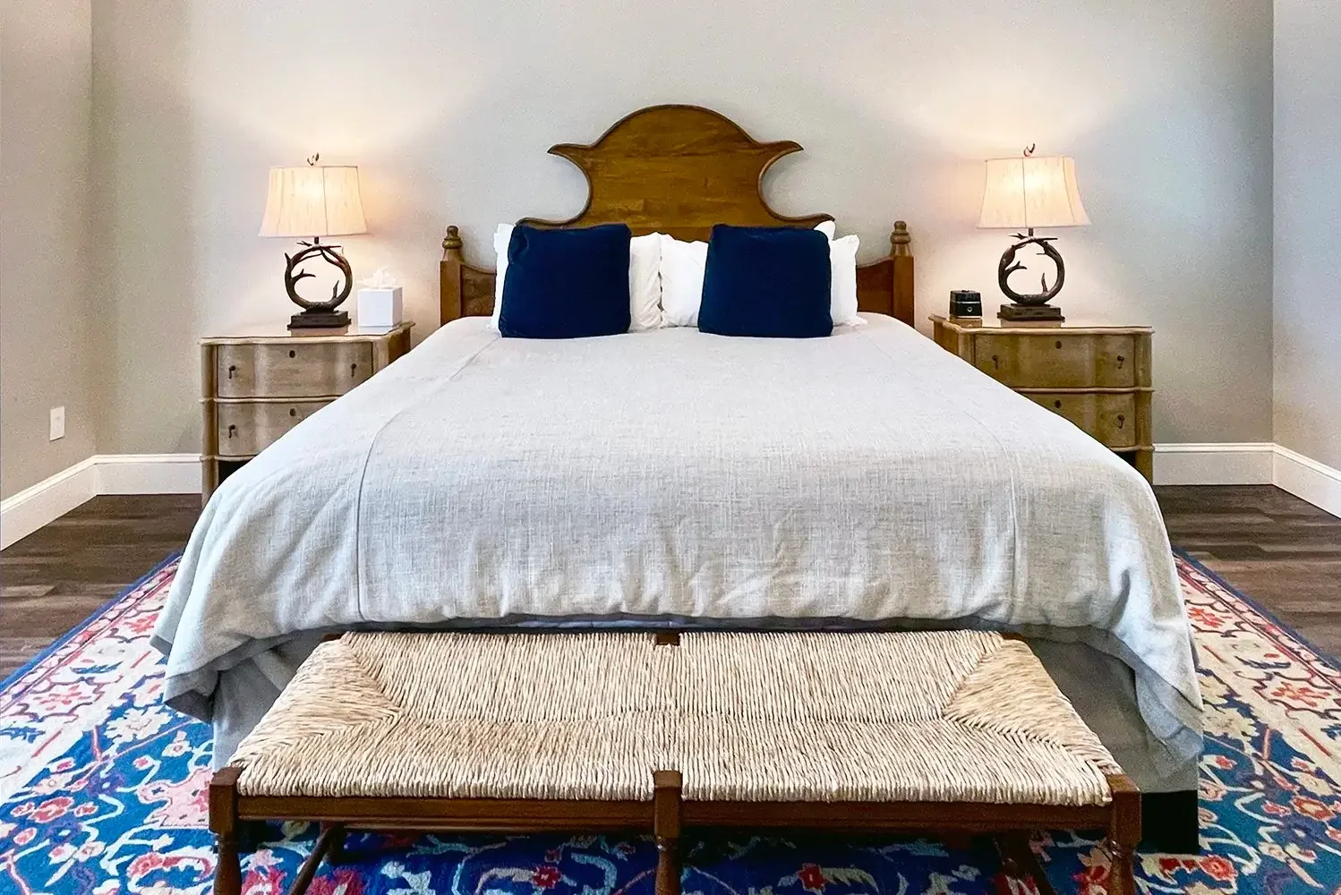 A neatly made bed with a wooden headboard, two dark blue pillows, and white bedding, flanked by two nightstands with lamps, set in a bedroom with hardwood floors and a decorative rug.