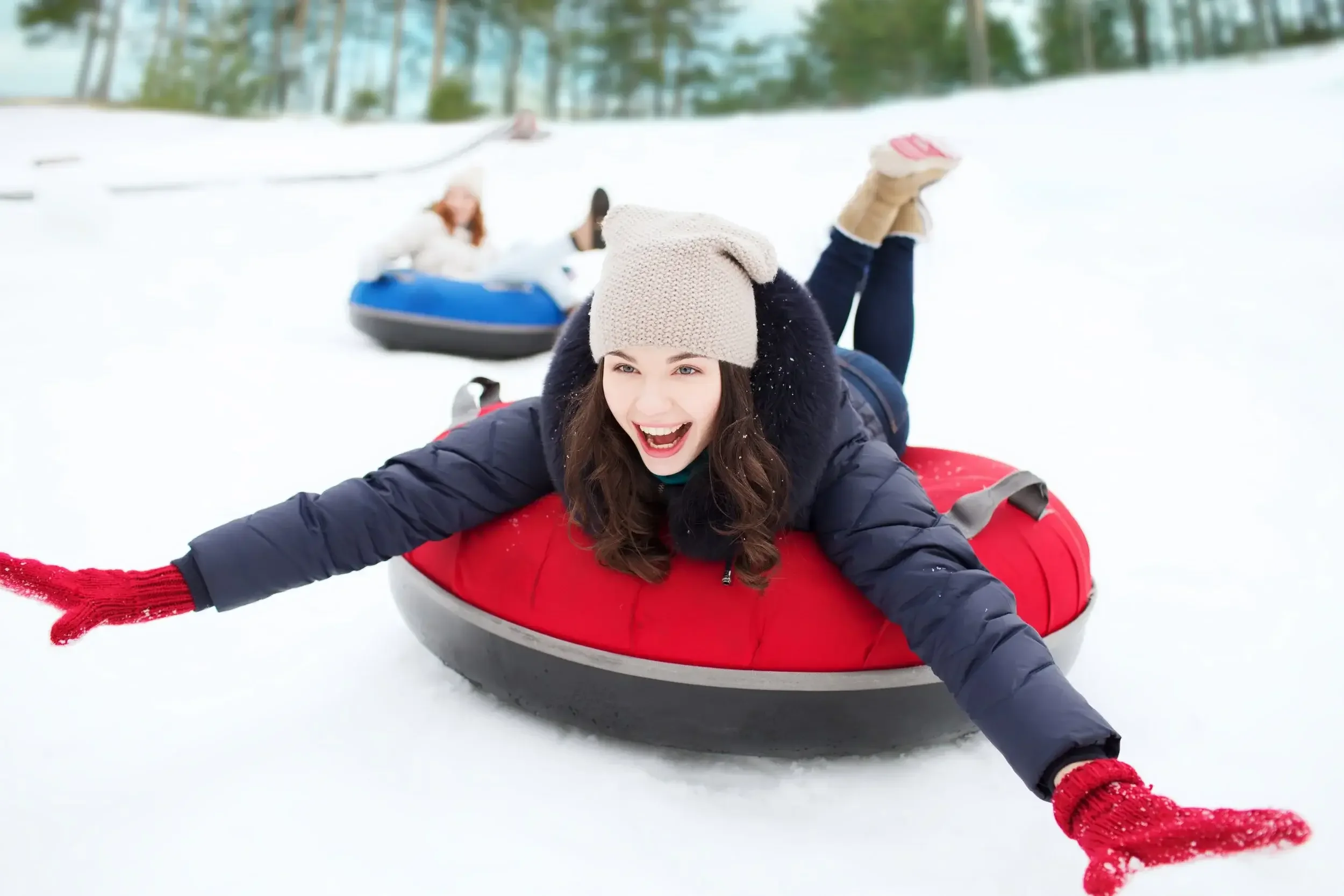 Young woman in winter clothes sledding down a snowy hill on a red snow tube, smiling and reaching out with her arms.