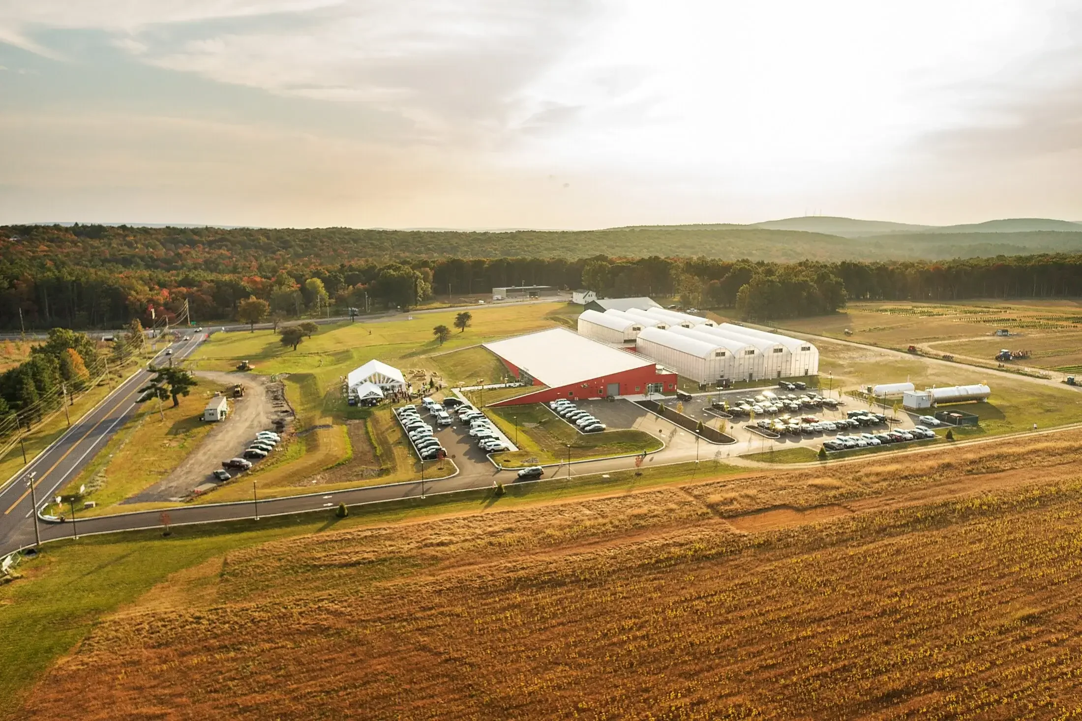 An aerial view of a large agricultural greenhouse complex with multiple white and red greenhouses, surrounded by parking lots, roads, and fields, with a backdrop of wooded hills and open farmland.