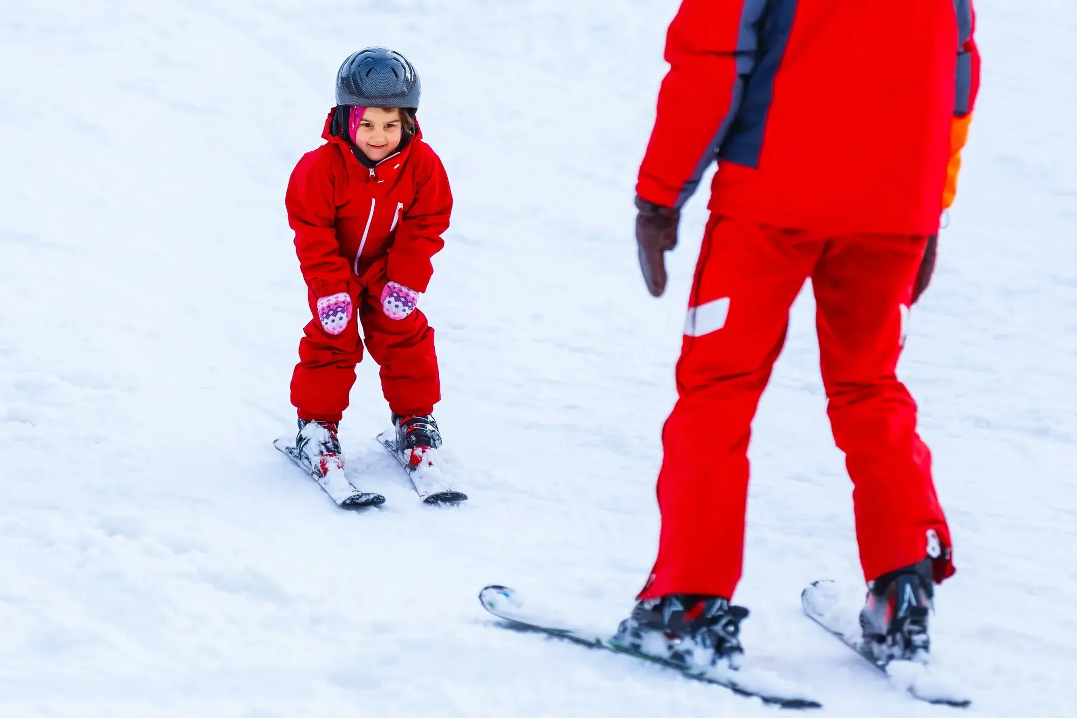 A young girl in a red snowsuit and pink gloves learning to ski from an adult in red ski gear on snowy terrain.