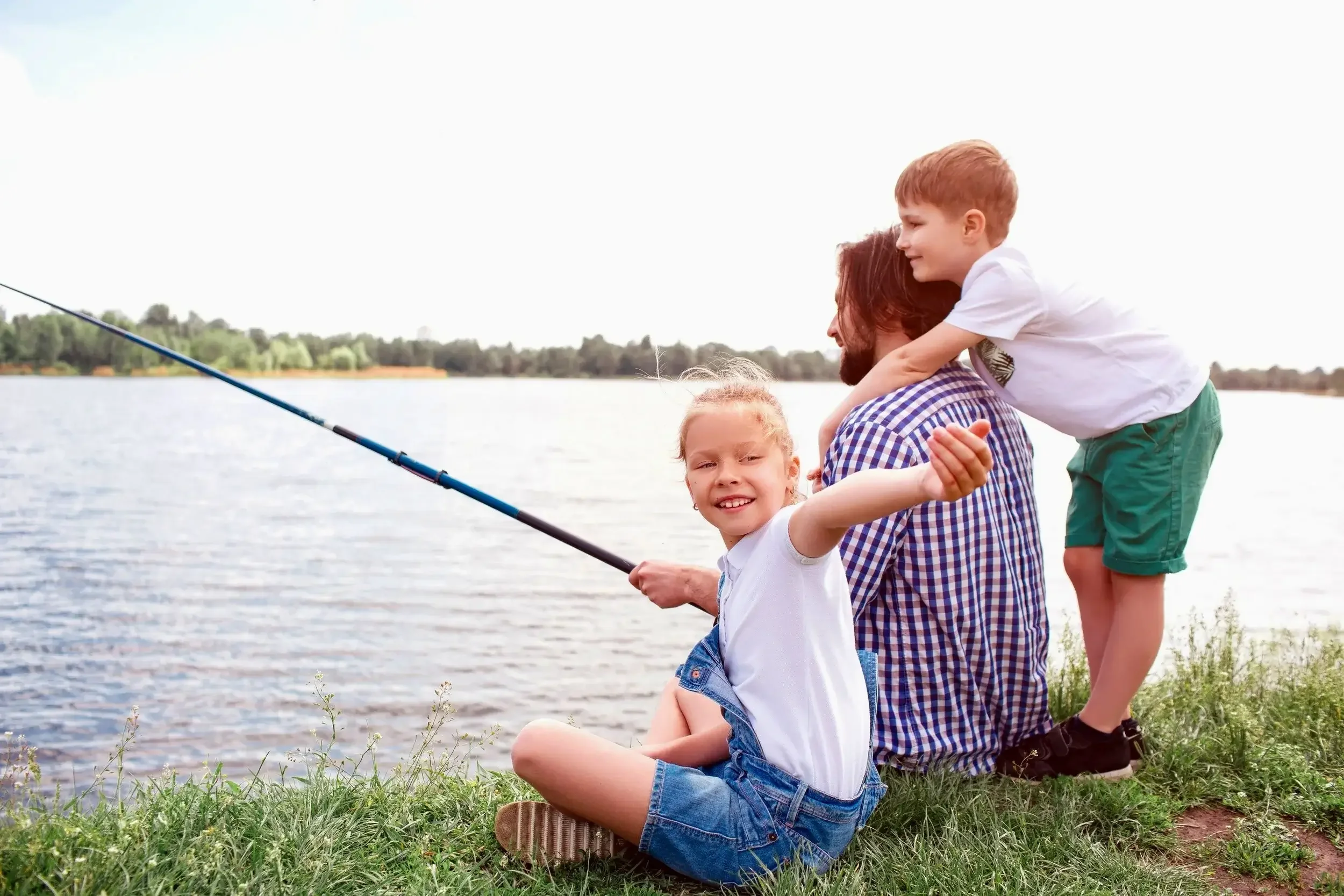 Dad sitting on bank of lake fishing with young son and daughter