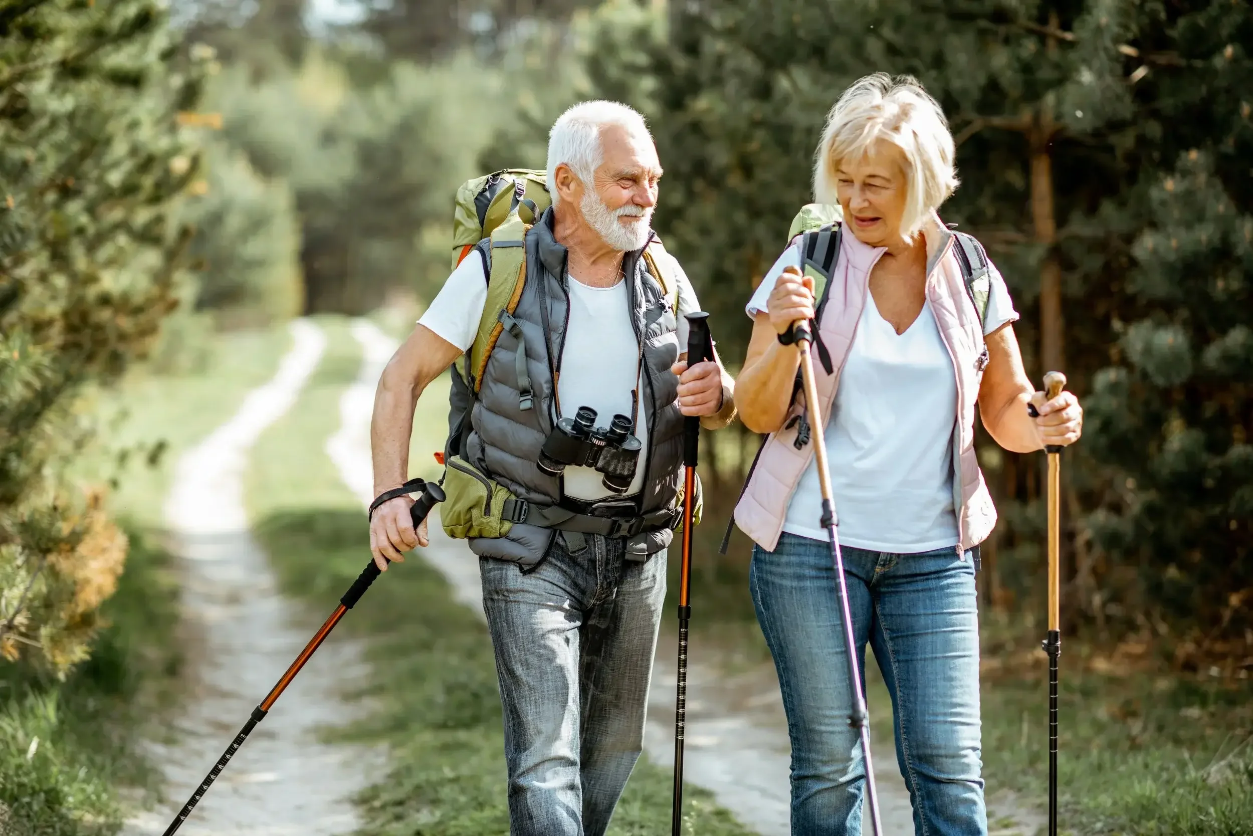 Senior couple hiking on a forest trail with backpacks and trekking poles.