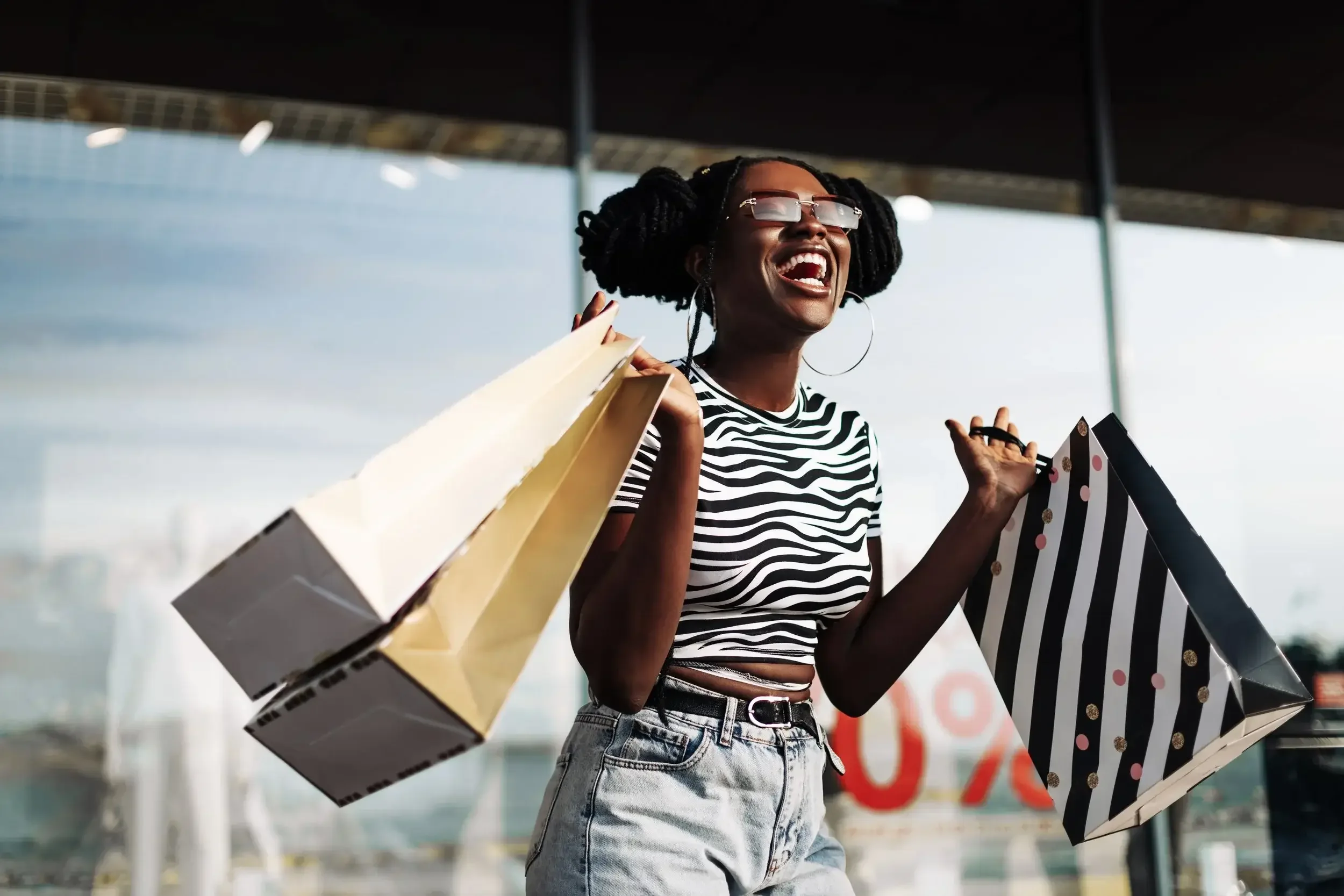 A young woman with dark skin, in a black and white striped crop top and high-waisted denim shorts, is smiling and carrying shopping bags while standing outdoors at a shopping mall.