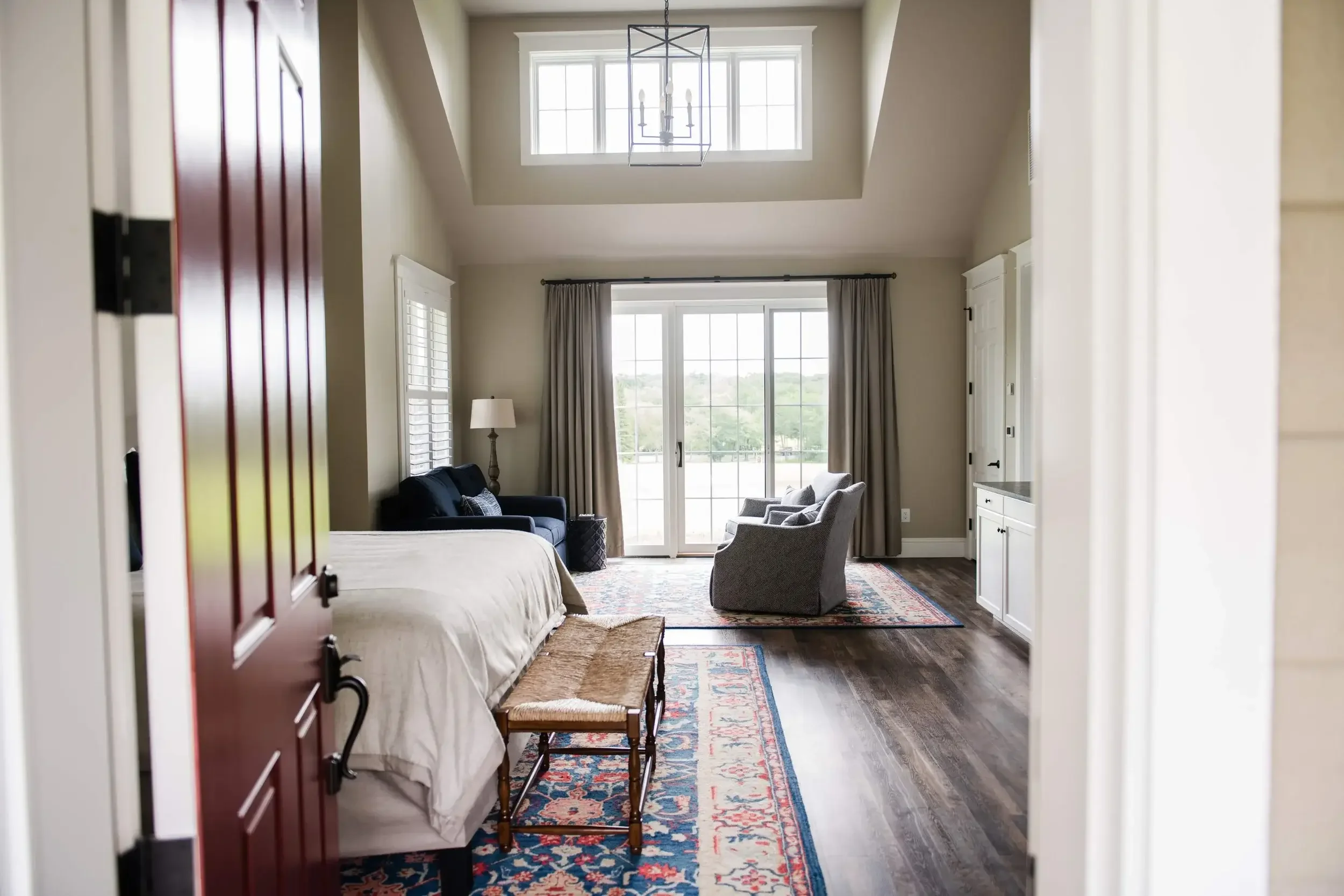 A modern bedroom with high ceilings, large windows, a bed with white linens, a black sofa, two gray armchairs, and sliding glass doors leading to an outdoor area, with hardwood floors and a colorful rug.