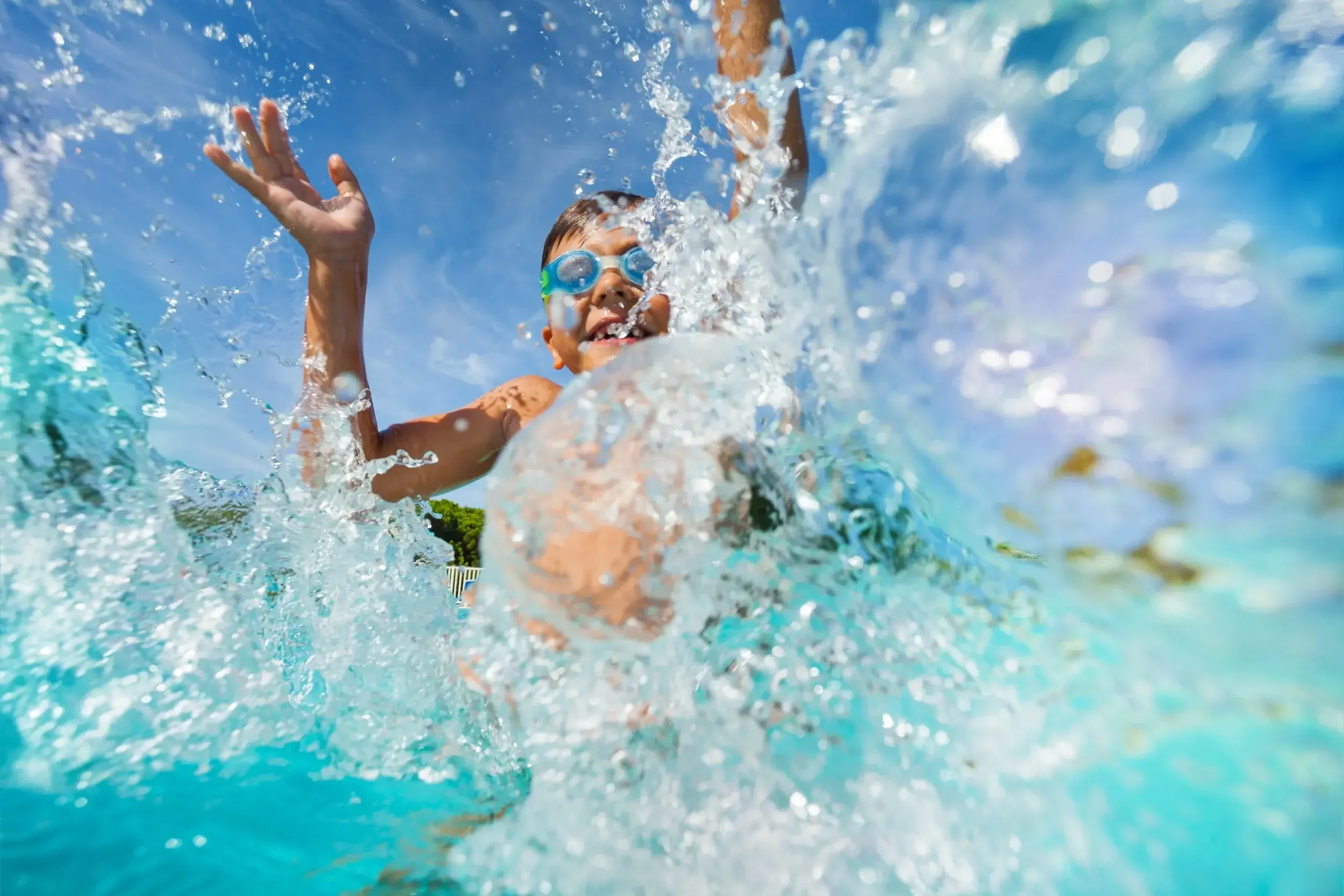 Child swimming in a pool with goggles, splashing water, under a bright blue sky.