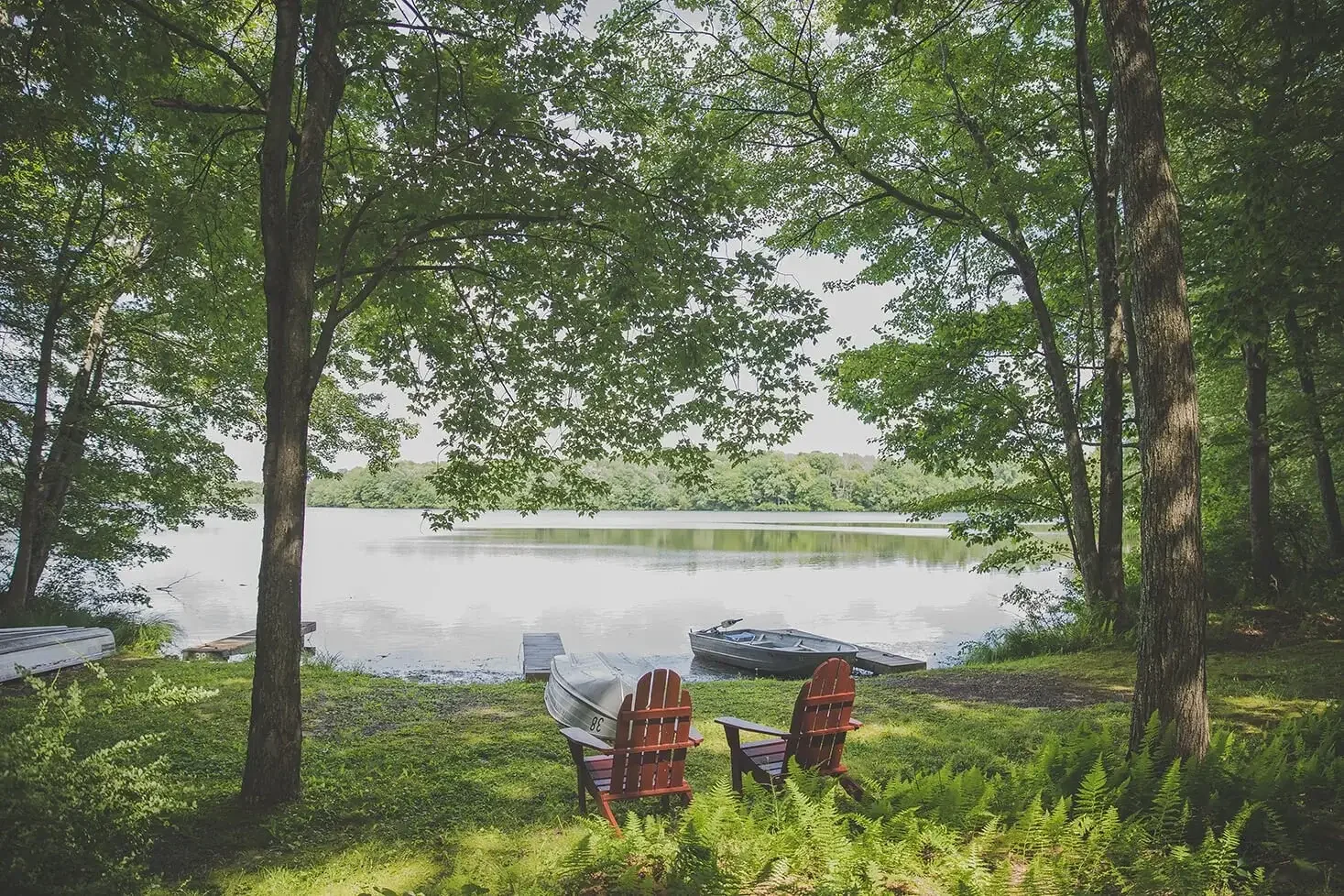 Two Adirondack chairs sit under the shade of tall trees, overlooking the lake with rowboats resting along the shoreline—a perfect spot for quiet relaxation.