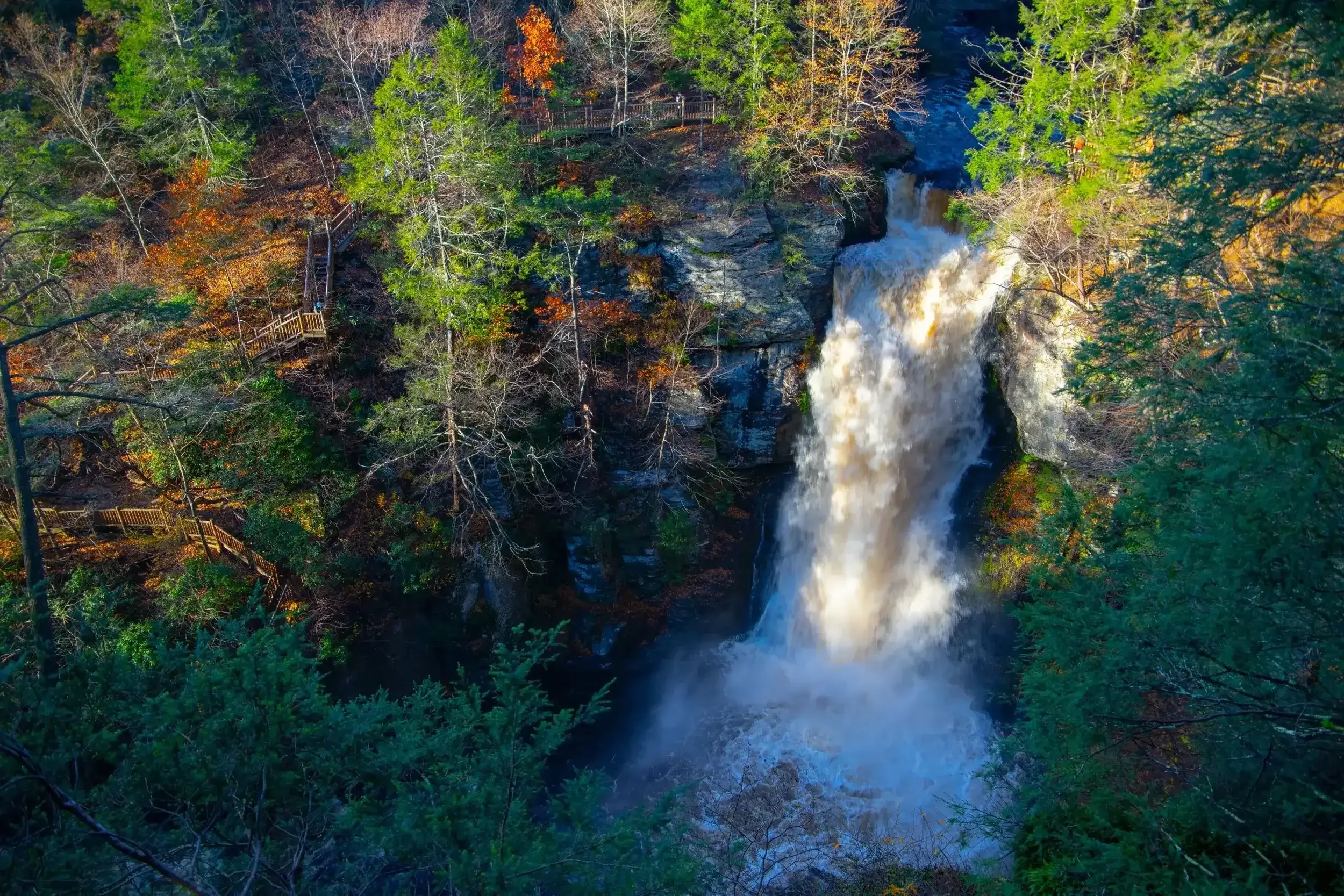 A waterfall cascading down a rocky cliff surrounded by trees with green and fall-colored leaves, with a wooden walkway running through the forest.