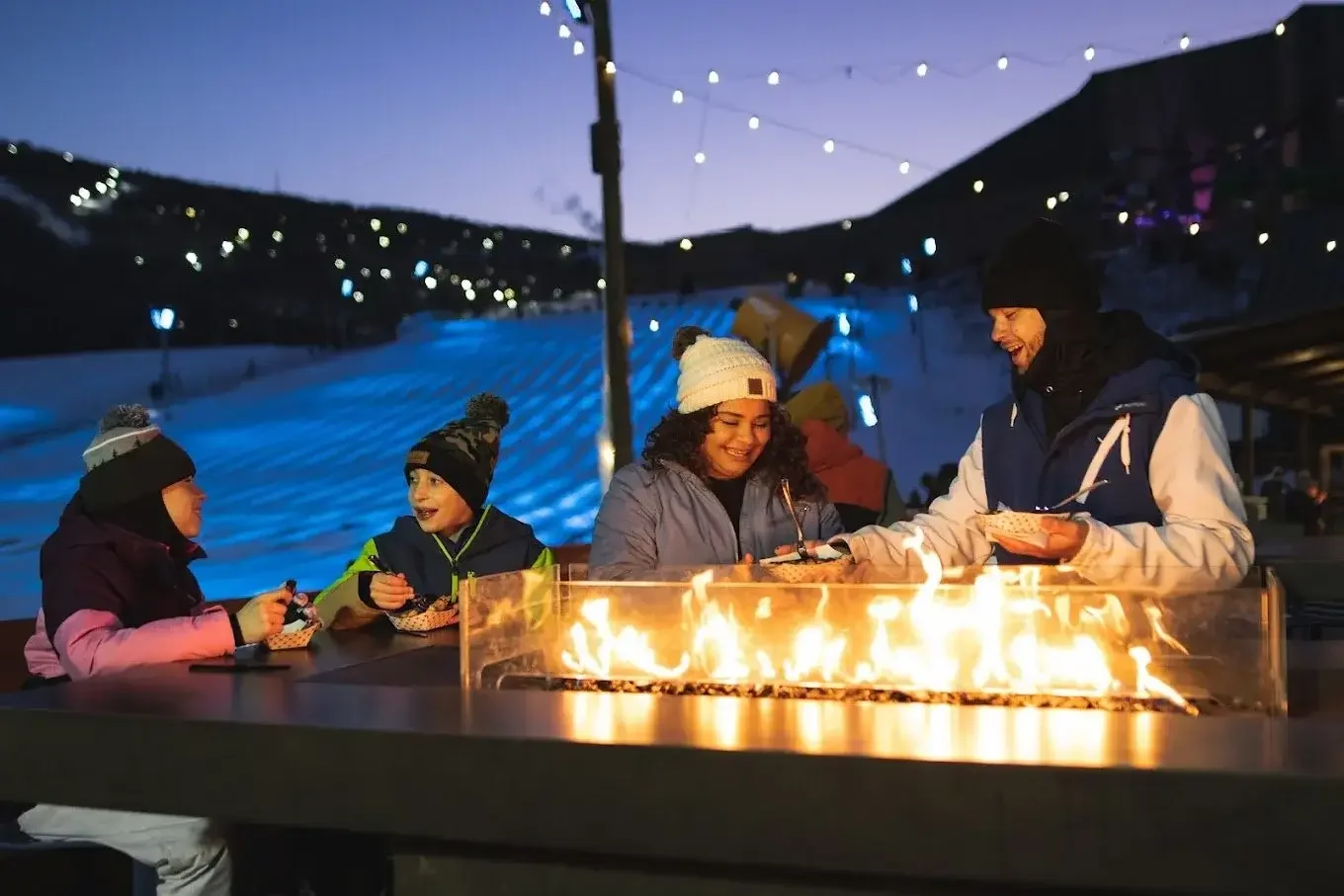 People at an outdoor fire pit enjoying food and conversation during winter, with snow and hillside lights in the background at dusk.