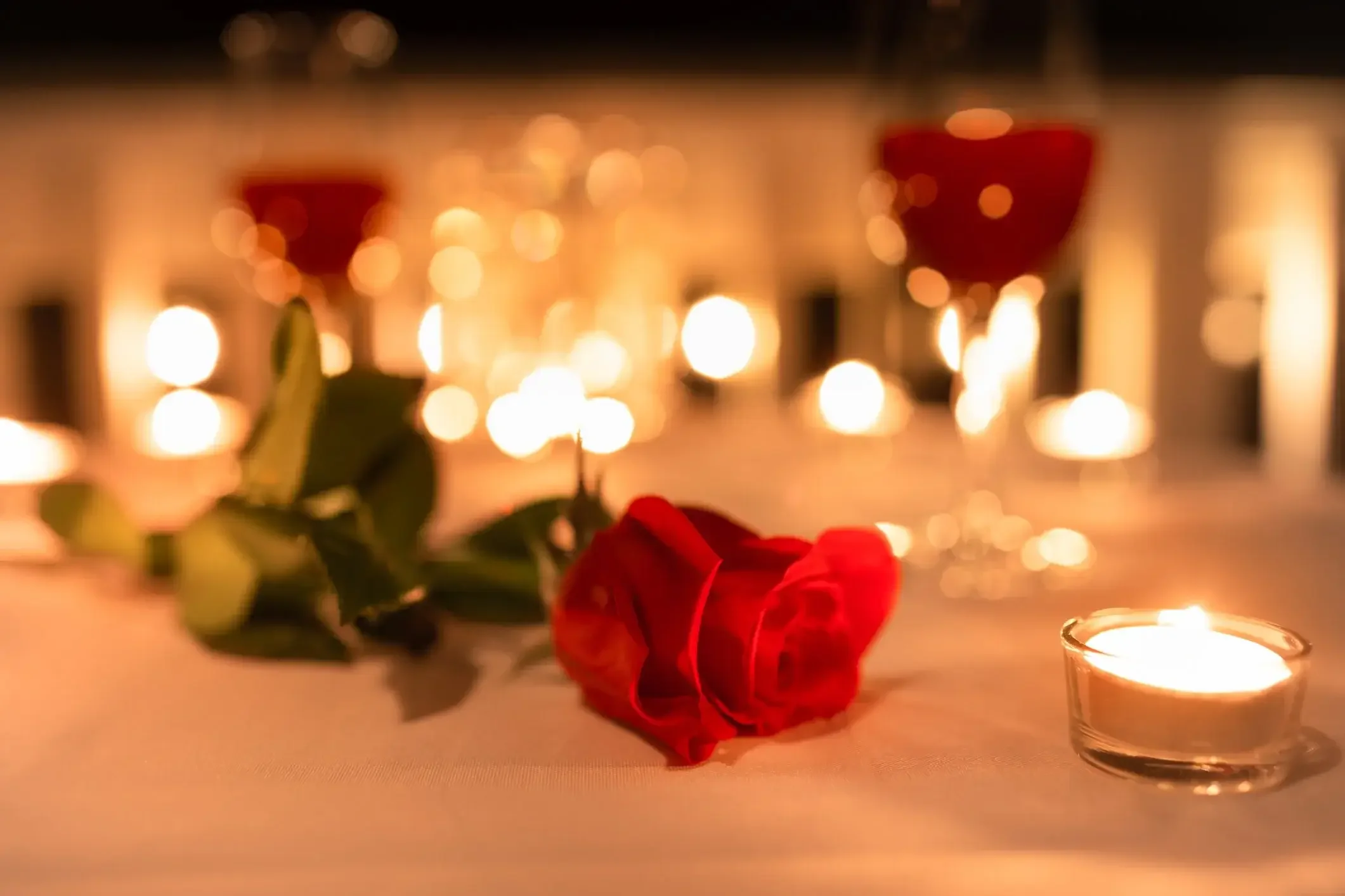 A single red rose with green leaves lying on a table, surrounded by lit candles and glasses of red wine, with a blurred background of candlelight.