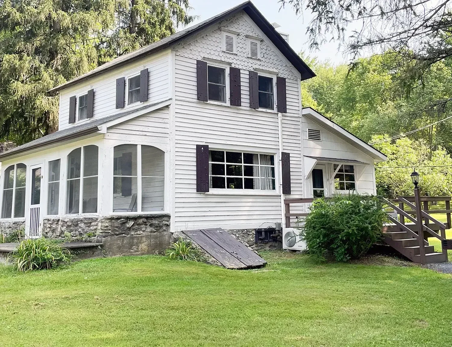 A two-story white house with black shutters, a stone foundation, and a screened porch. There is a small ramp leading to the side entrance, and a set of stairs with a railing on the front. The house is surrounded by greenery and trees.