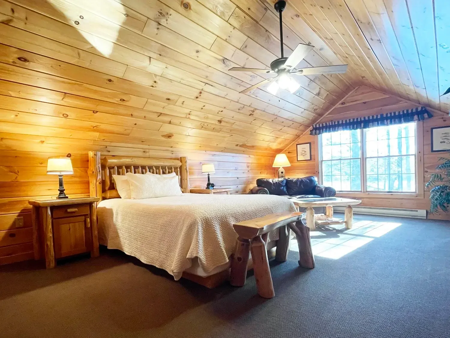 A cozy bedroom with wooden walls and a sloped ceiling, featuring a bed with cream bedding, two bedside lamps, a black leather sofa, a wooden coffee table, and a large window with a valance, allowing natural light into the room.