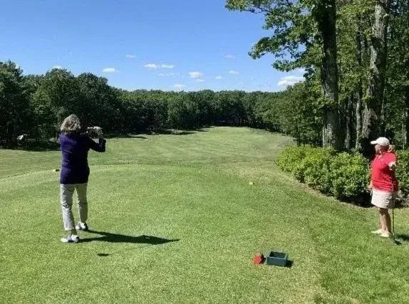 Two women on a golf course, one taking a shot and the other observing, with golf equipment and a ball near the tee box, surrounded by trees under a blue sky.