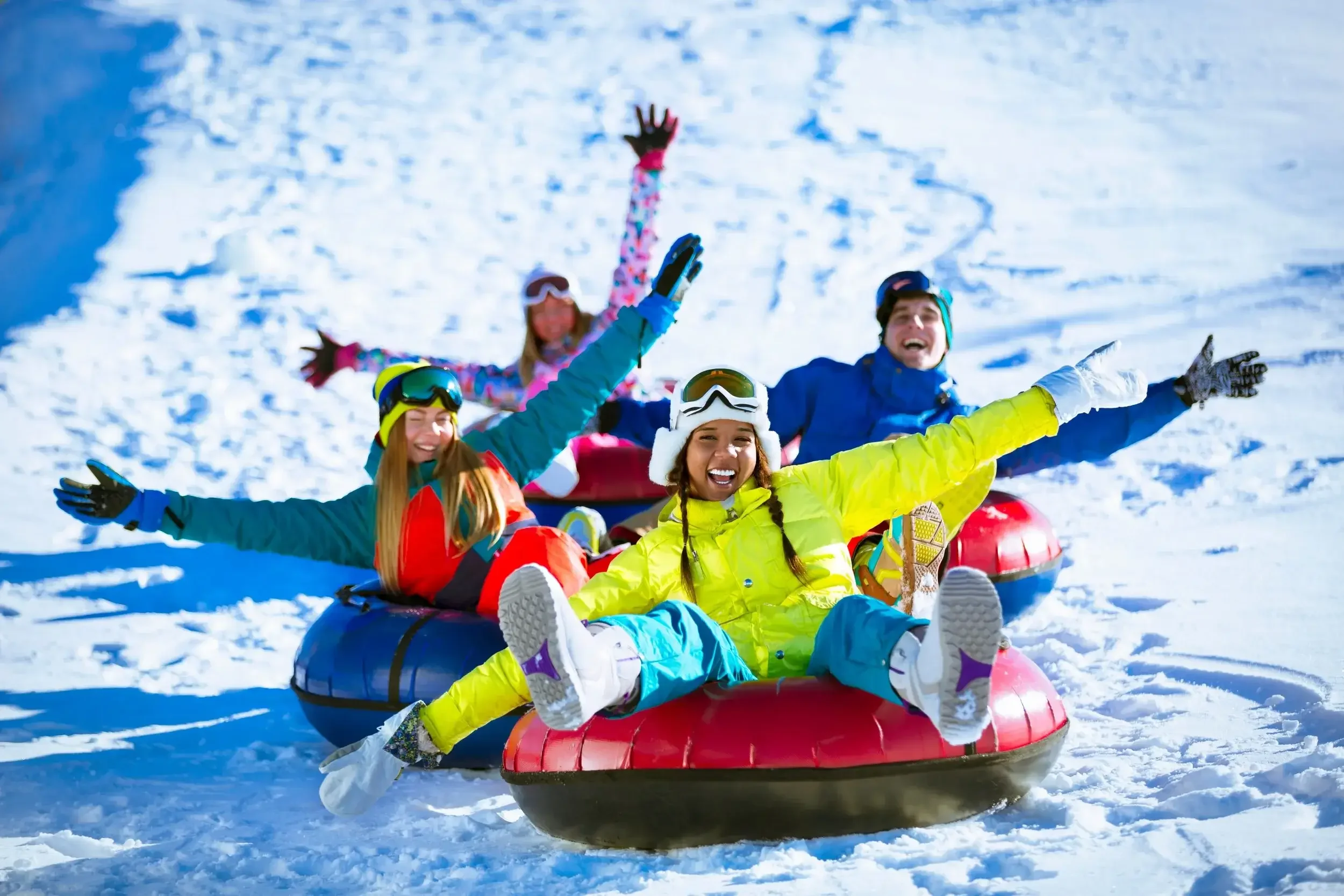 Group of people in colorful winter clothing sledding down a snowy hill, smiling and enjoying the activity.