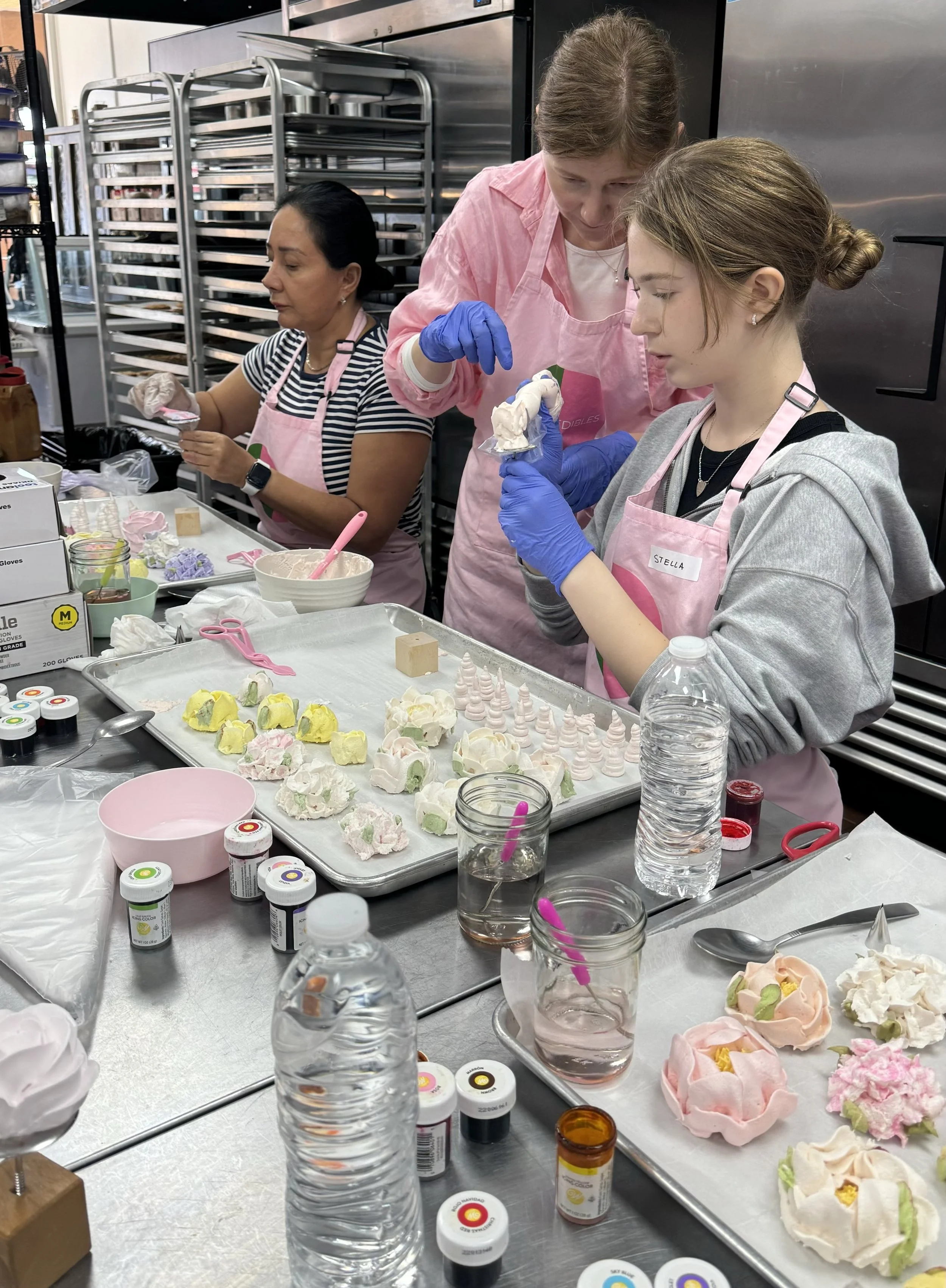 Three people decorating meringue flowers in a bakery, wearing aprons and gloves. Various baking supplies, including bowls and food coloring, are on tables.