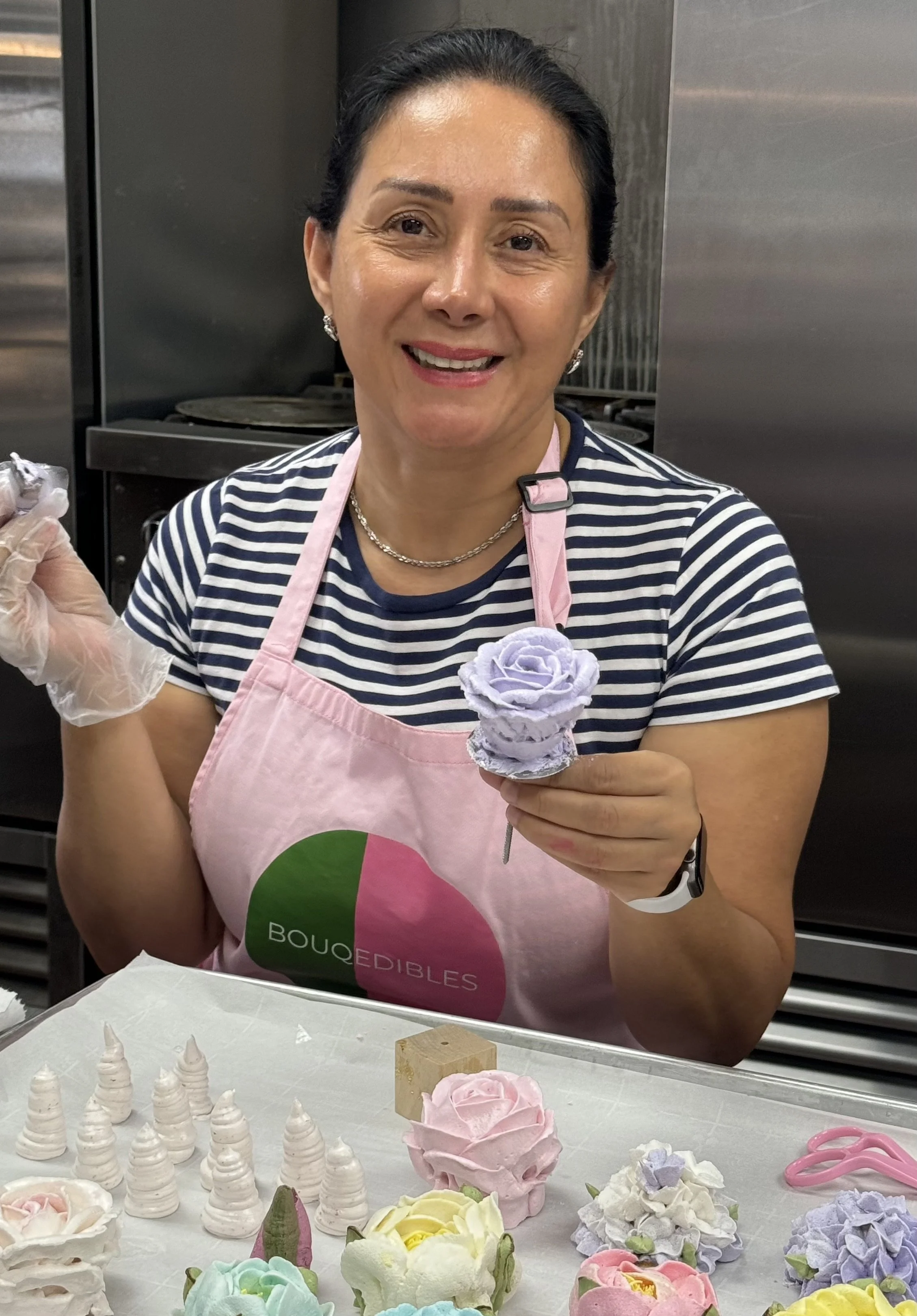 Woman in a striped shirt and pink apron holding a purple flower-shaped edible decoration, with more decorations on a table.