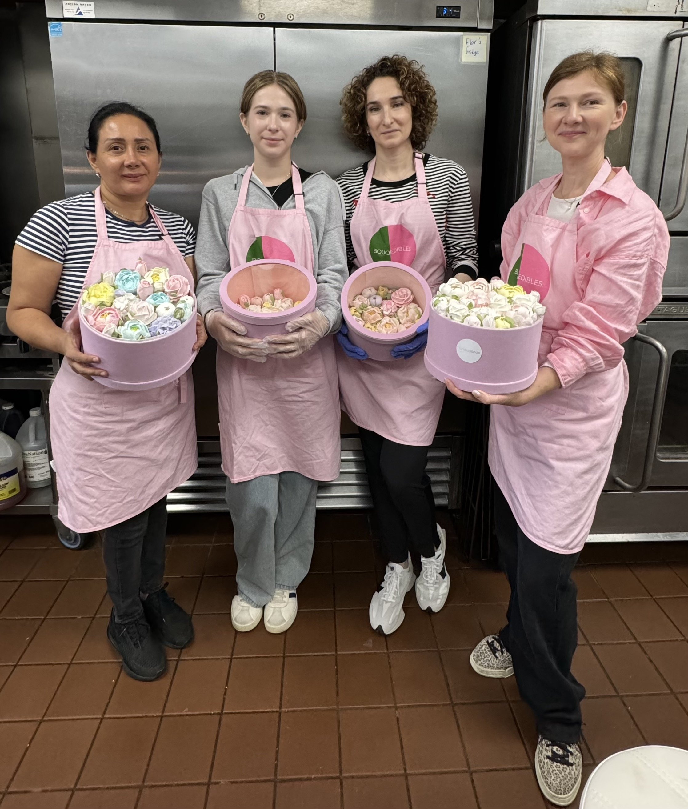 Four women wearing pink aprons and holding buckets of colorful meringues in a kitchen setting.