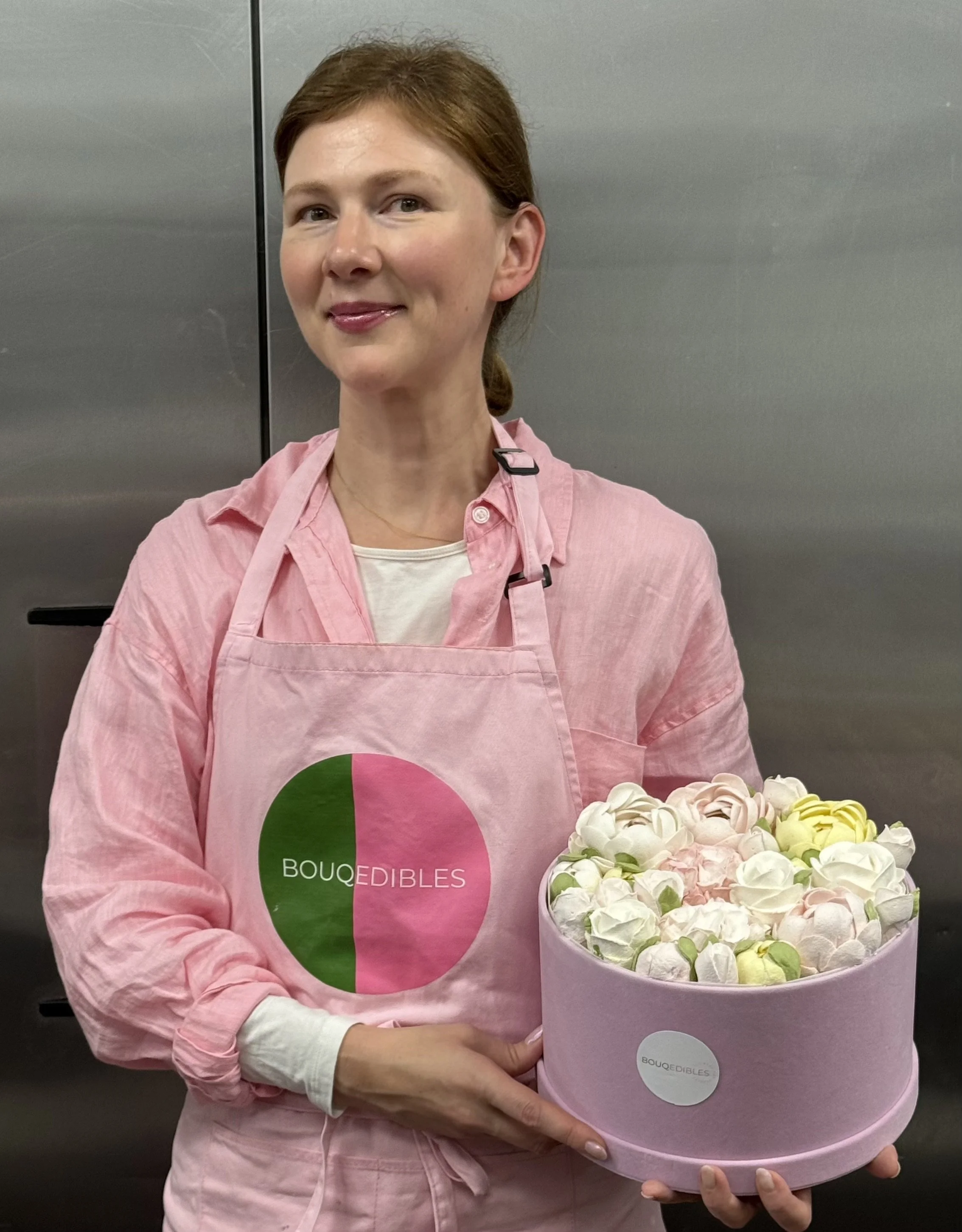 Woman in pink apron holding a flower arrangement in a pink round box.