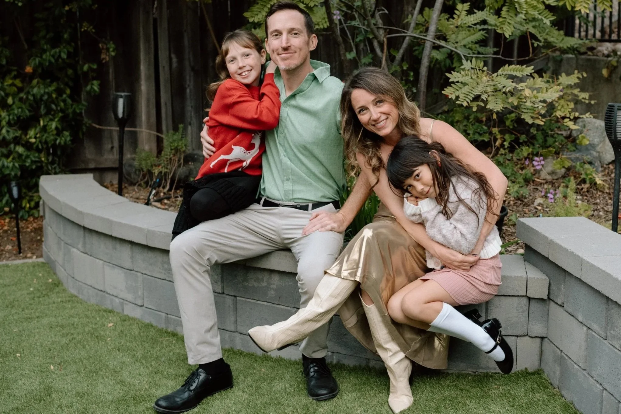 Family of four sitting outdoors on a curved stone wall, smiling and hugging each other, with trees and garden in the background.