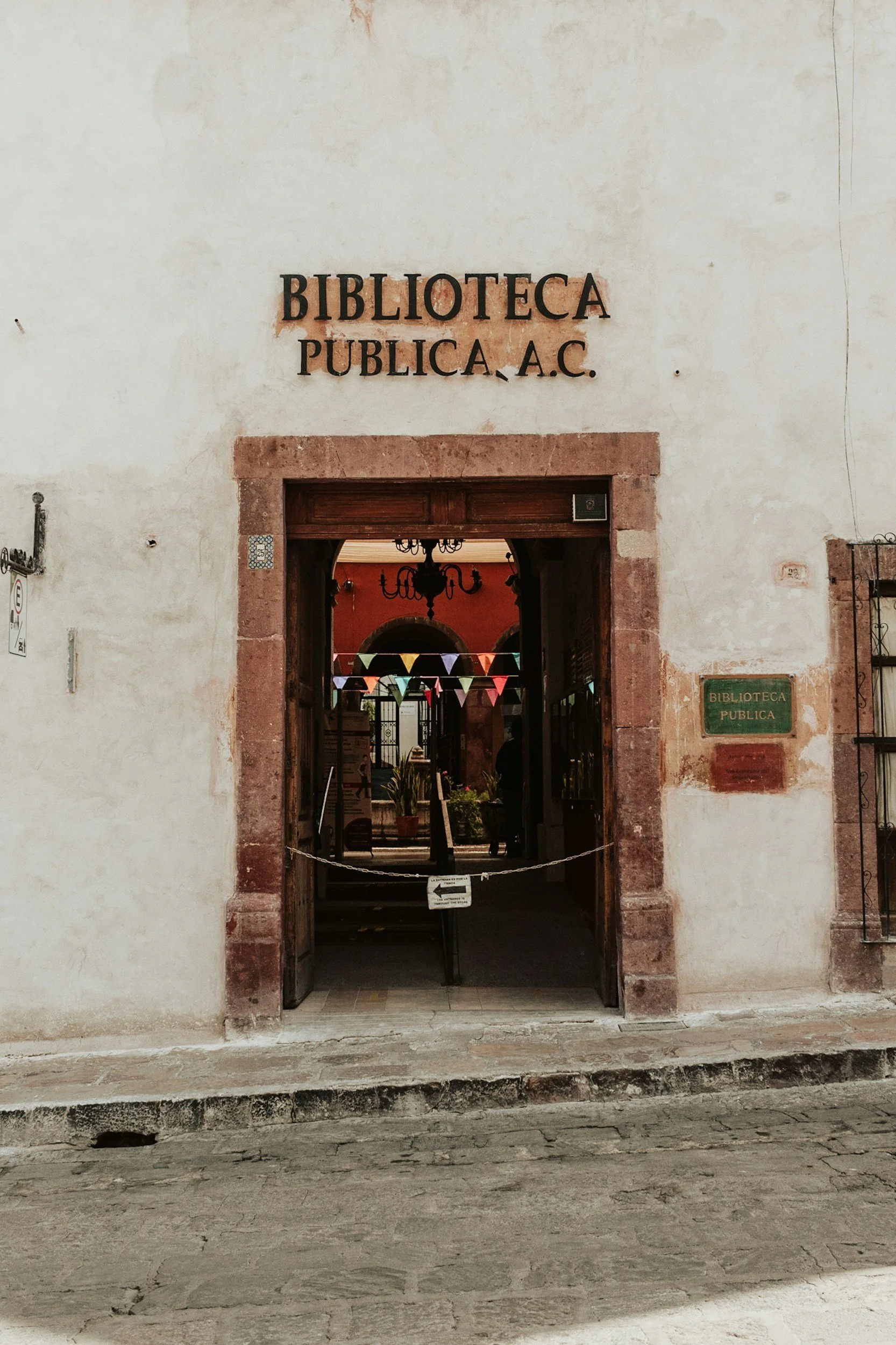 Entrance to a public library building with a sign that reads 'Biblioteca Publica A.C.' and a doorway decorated with colorful bunting flags inside.