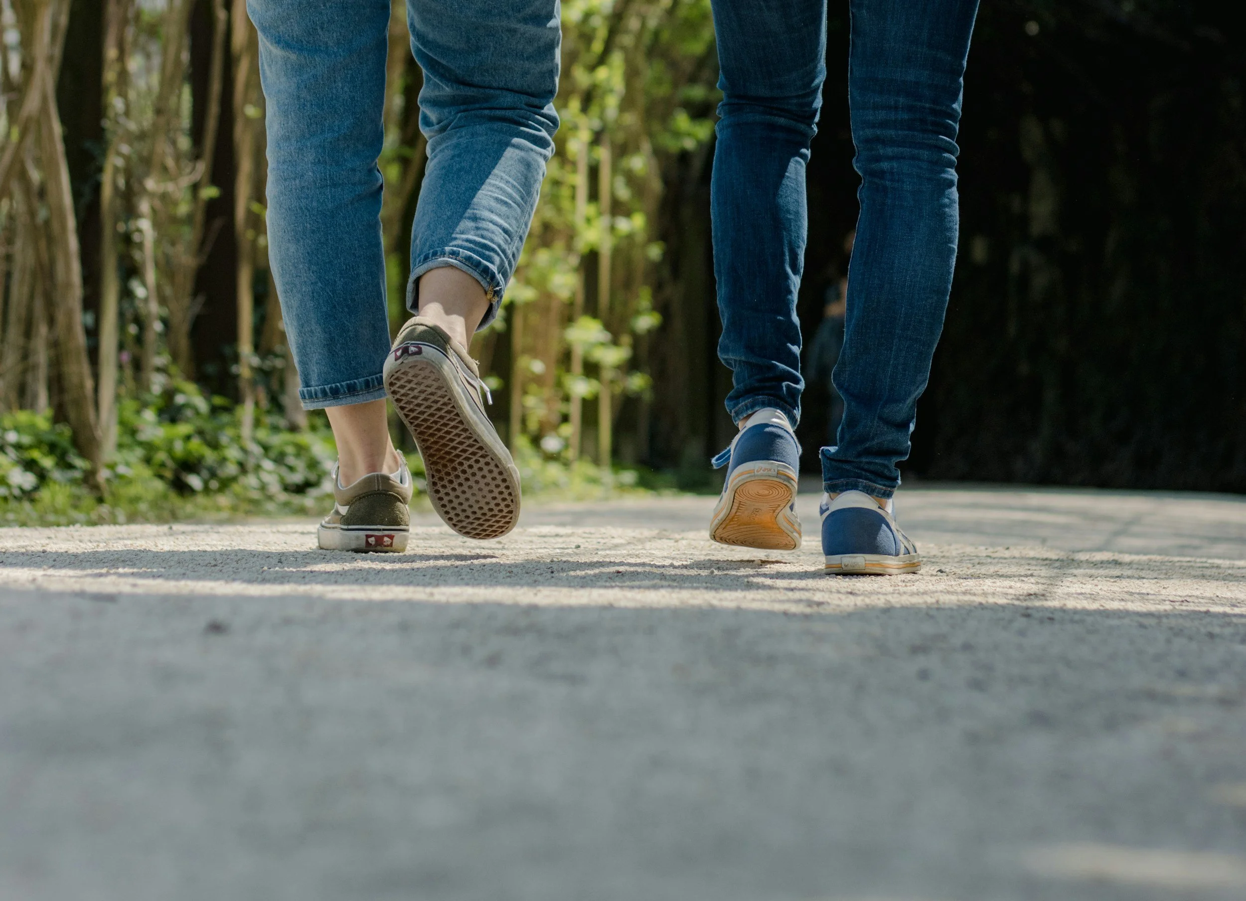 Photograph of two people walking outdoors along a path, focusing on their legs and feet, with a background of trees and greenery.