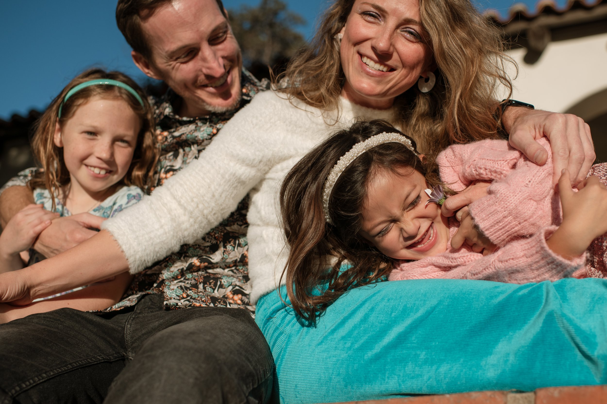 Family of four, including two young girls, a woman, a man, lying closely together outside under clear blue sky, smiling and laughing.