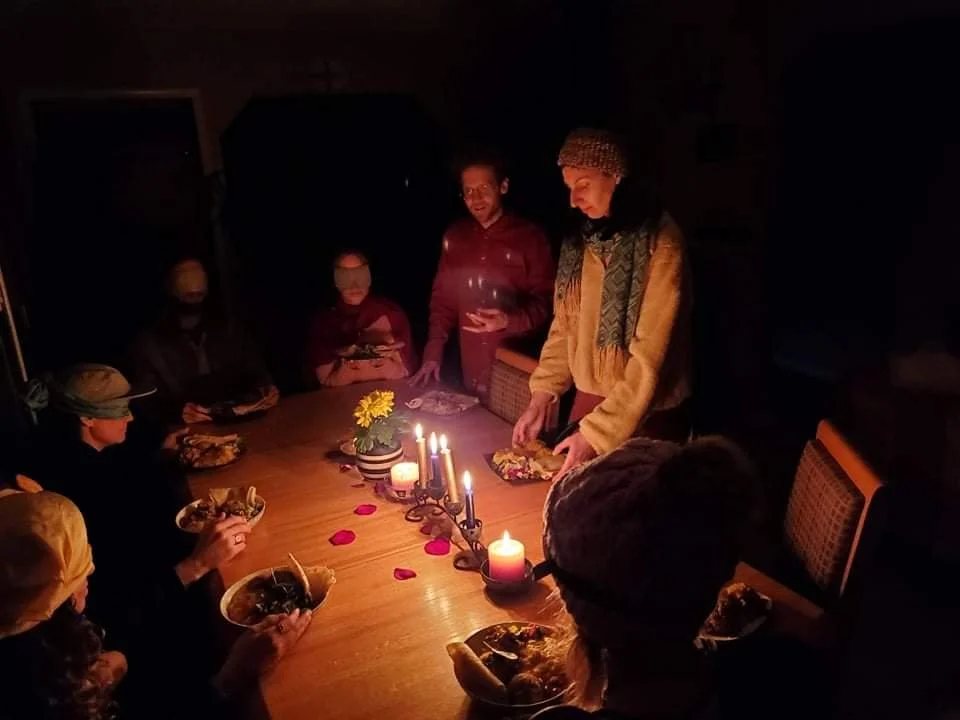 A family gathered around a dining table for a meal in a dimly lit room, illuminated by candles. The table has food, a candle centerpiece, and rose petals, with some family members wearing hats or scarves.