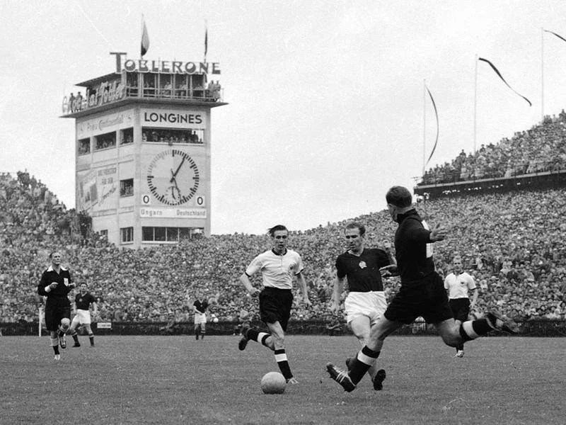 Players clash at the "Miracle at the Bern" in 1954 with recognizable global Swiss brands displayed on a clocktower in the background.