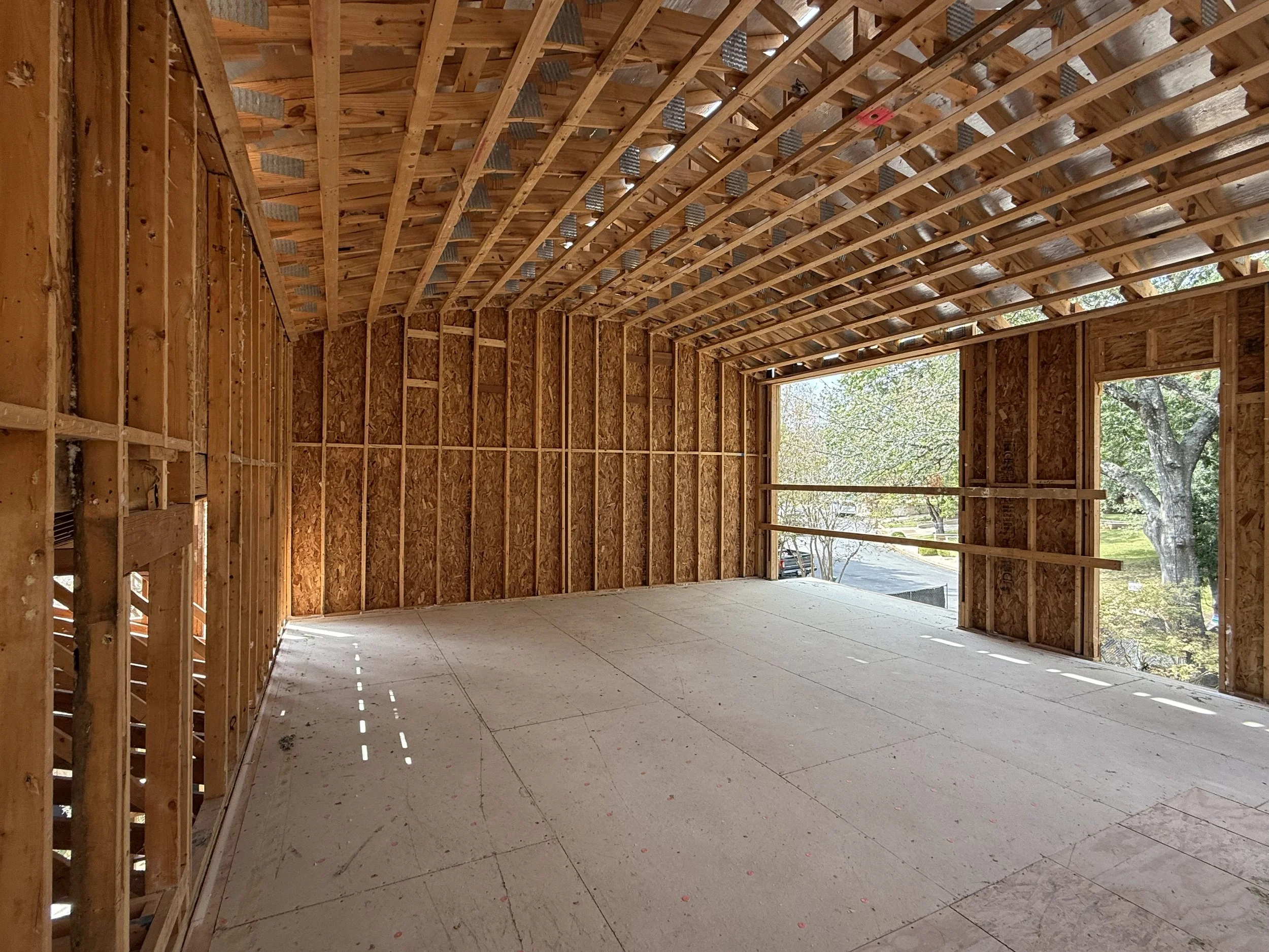 Interior view of a house under construction with wooden framing, exposed roof trusses, and large open windows.