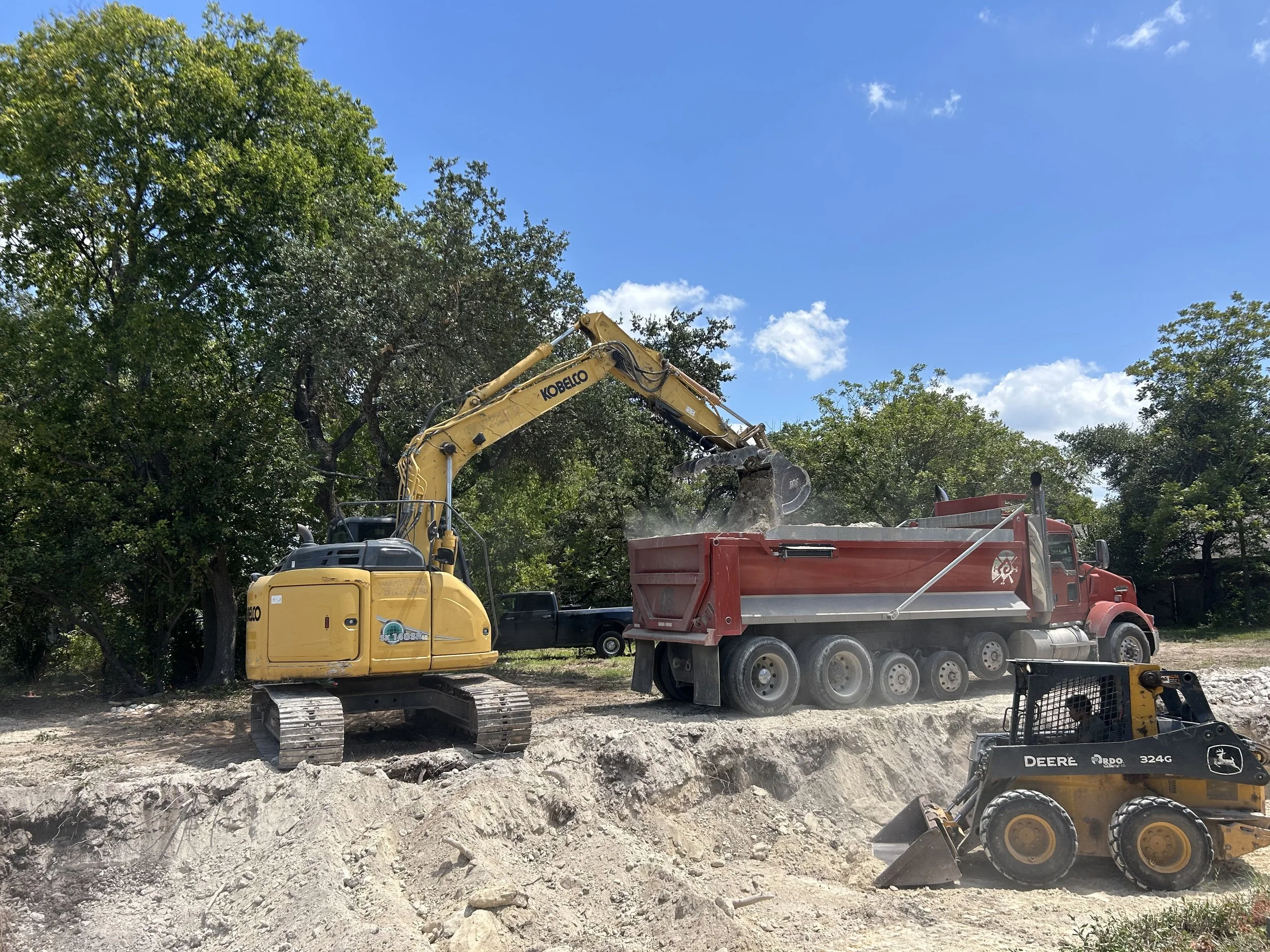 Construction site with a yellow Kobelco excavator loading dirt into a large red dump truck, a John Deere skid steer loader nearby, and trees in the background under a blue sky.