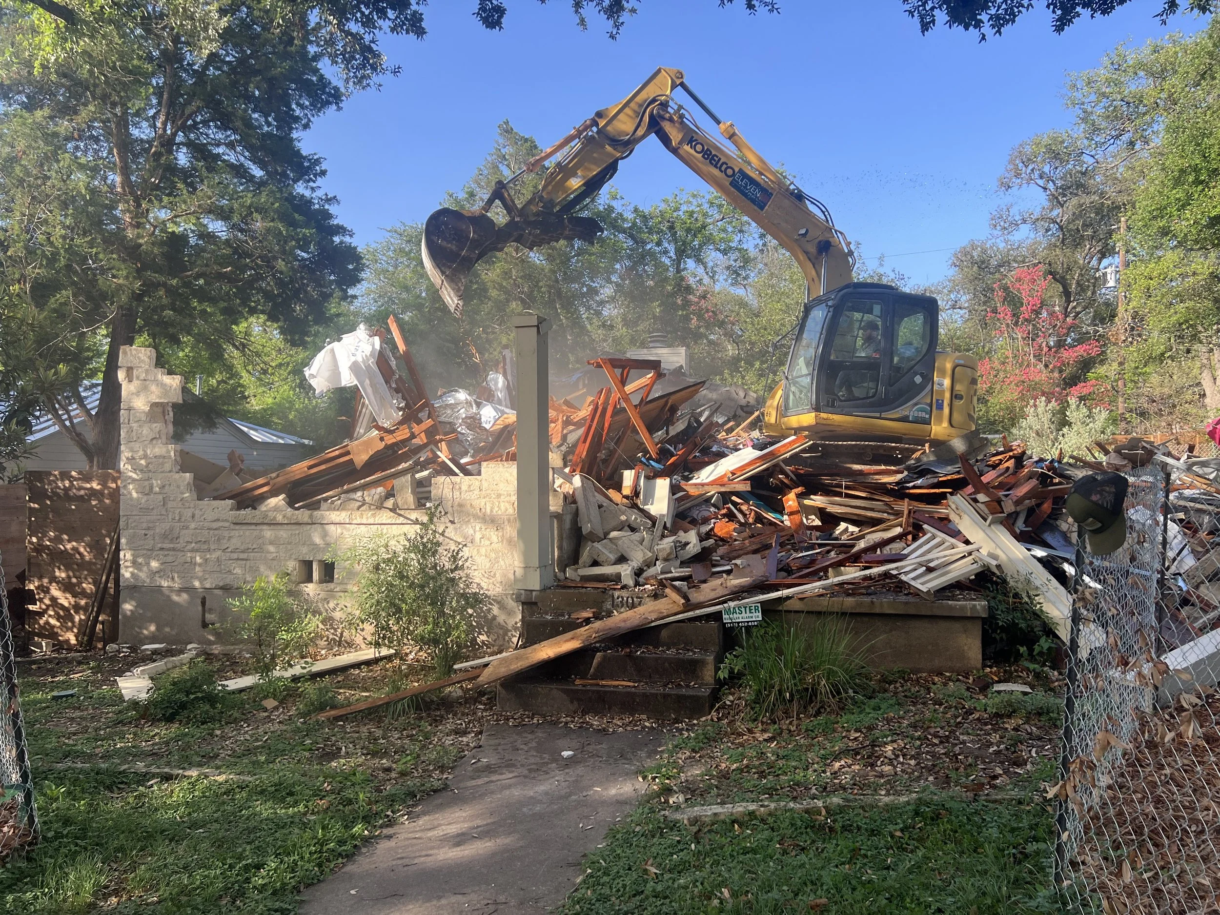 A yellow excavator demolishing a house, with debris and broken wood scattered around on a partly cloudy day with trees in the background.
