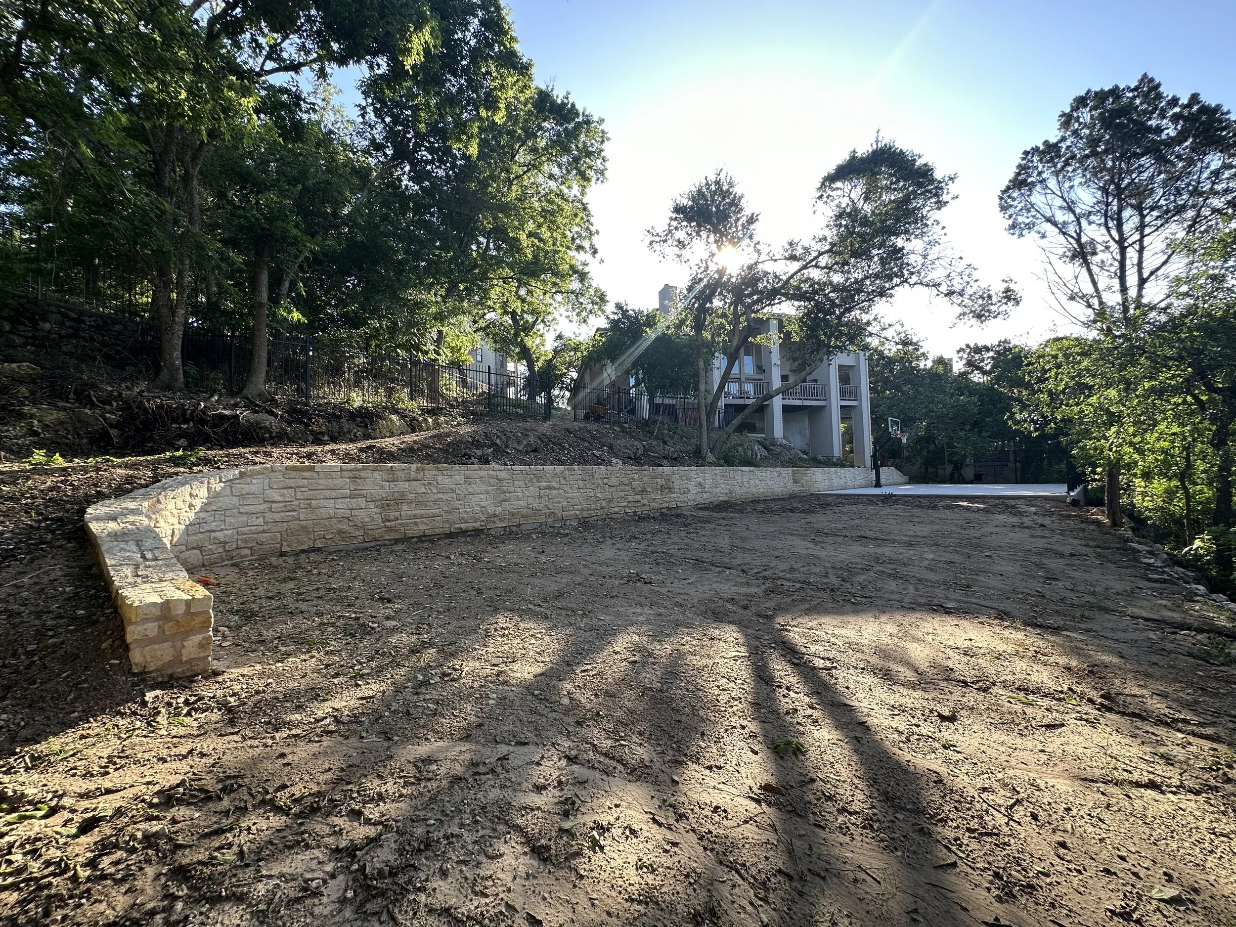A backyard with a dirt driveway, a stone retaining wall, tall trees, a house in the background, and sunlight filtering through the trees.