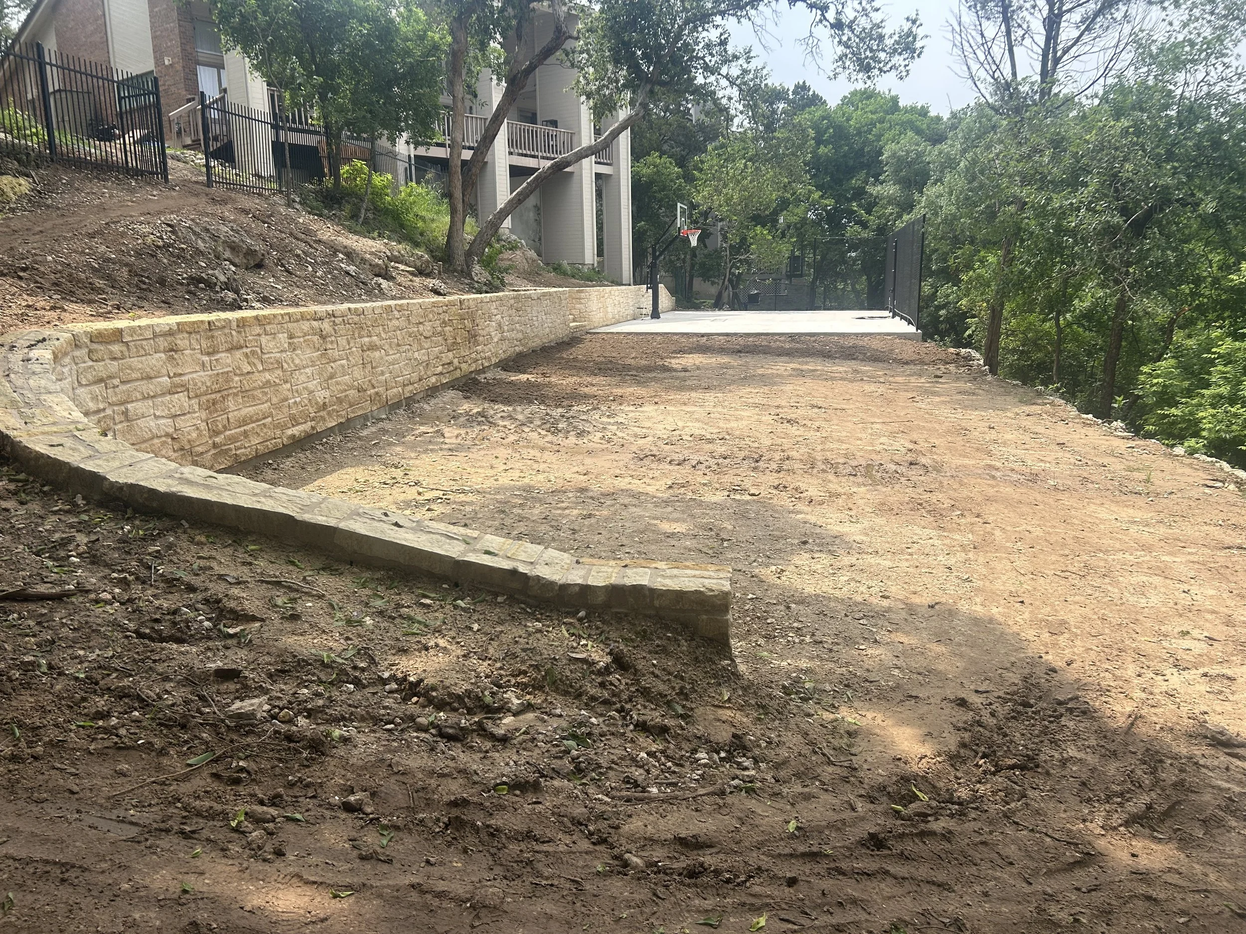 A backyard with a partially constructed stone retaining wall, dirt ground, in-ground basketball hoop, and a fenced sports court surrounded by trees and residential buildings.
