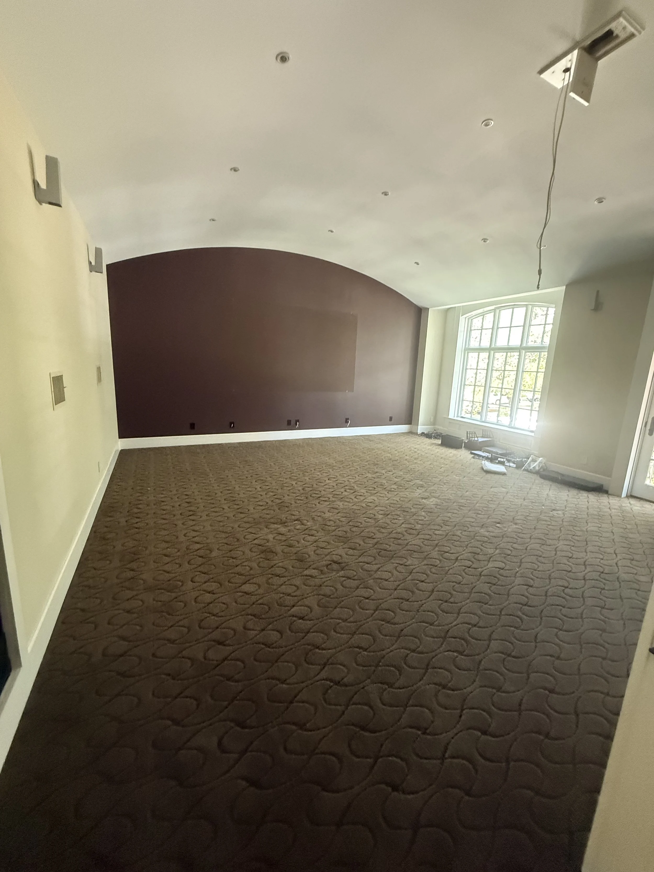Empty living room with brown patterned carpet, large window, and a dark accent wall.