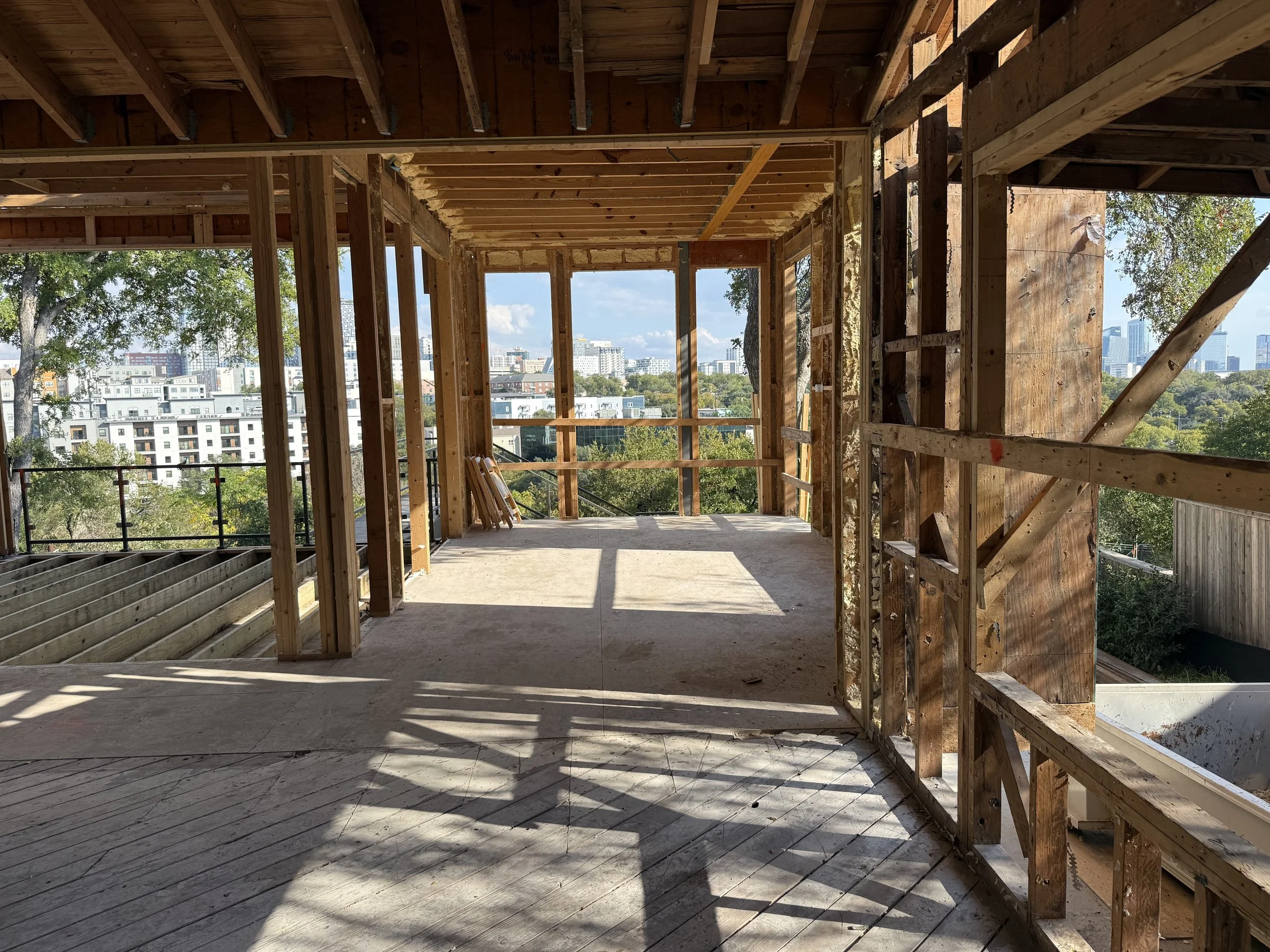 View of an under-construction building interior showing wooden framing, open walls, and a cityscape beyond the framed windows